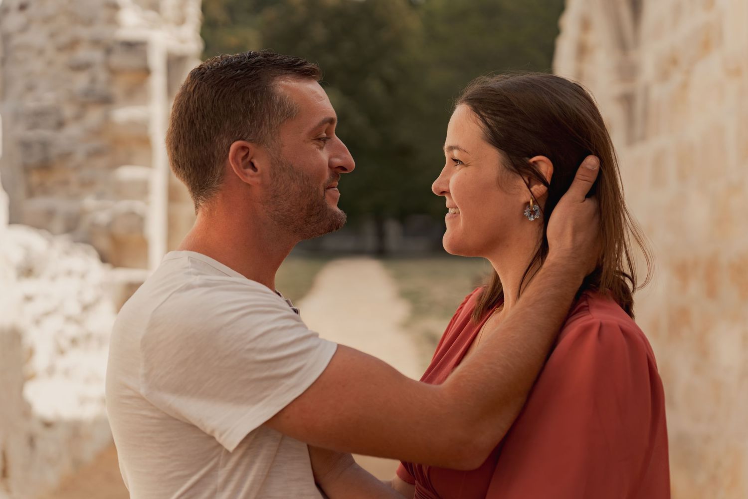 A couple shares a tender moment, man gently holding woman's face, smiling at each other near rustic stone walls.