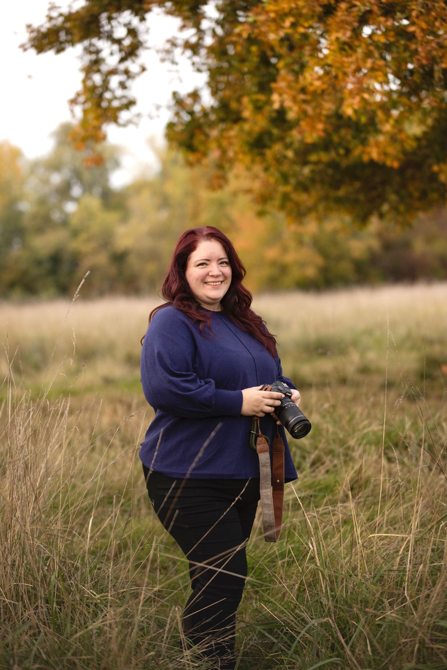 Woman with red hair holding a camera smiles in an autumn field with colorful foliage trees in background.