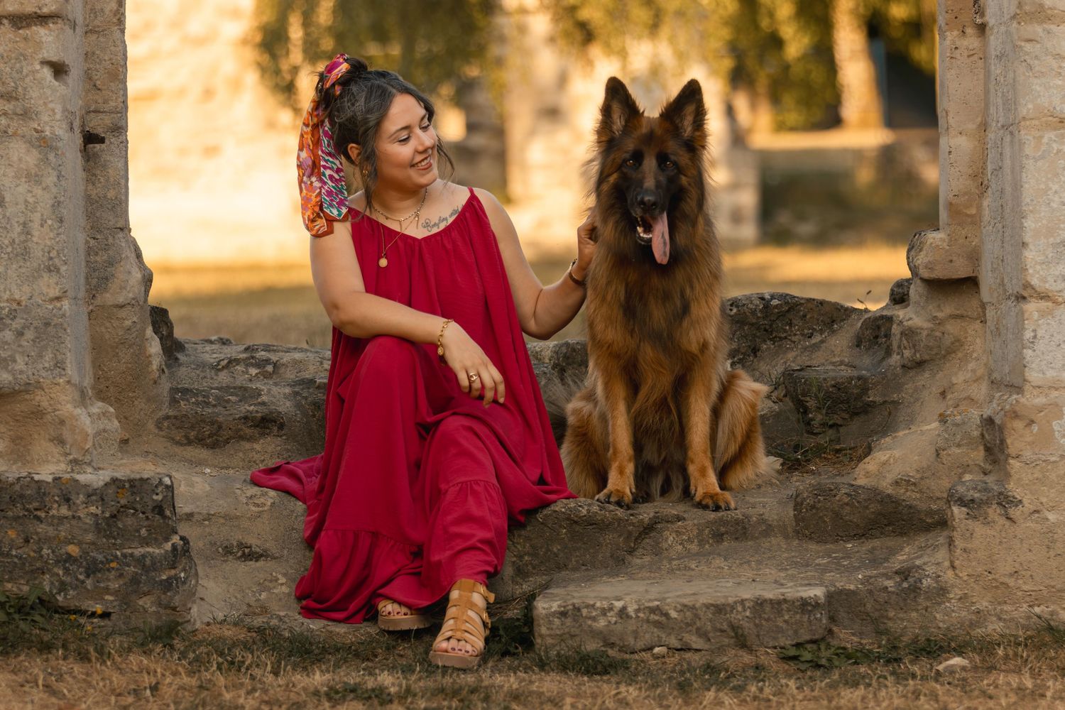 Woman in red dress sitting on stone steps with a German Shepherd dog in a sunlit outdoor setting.