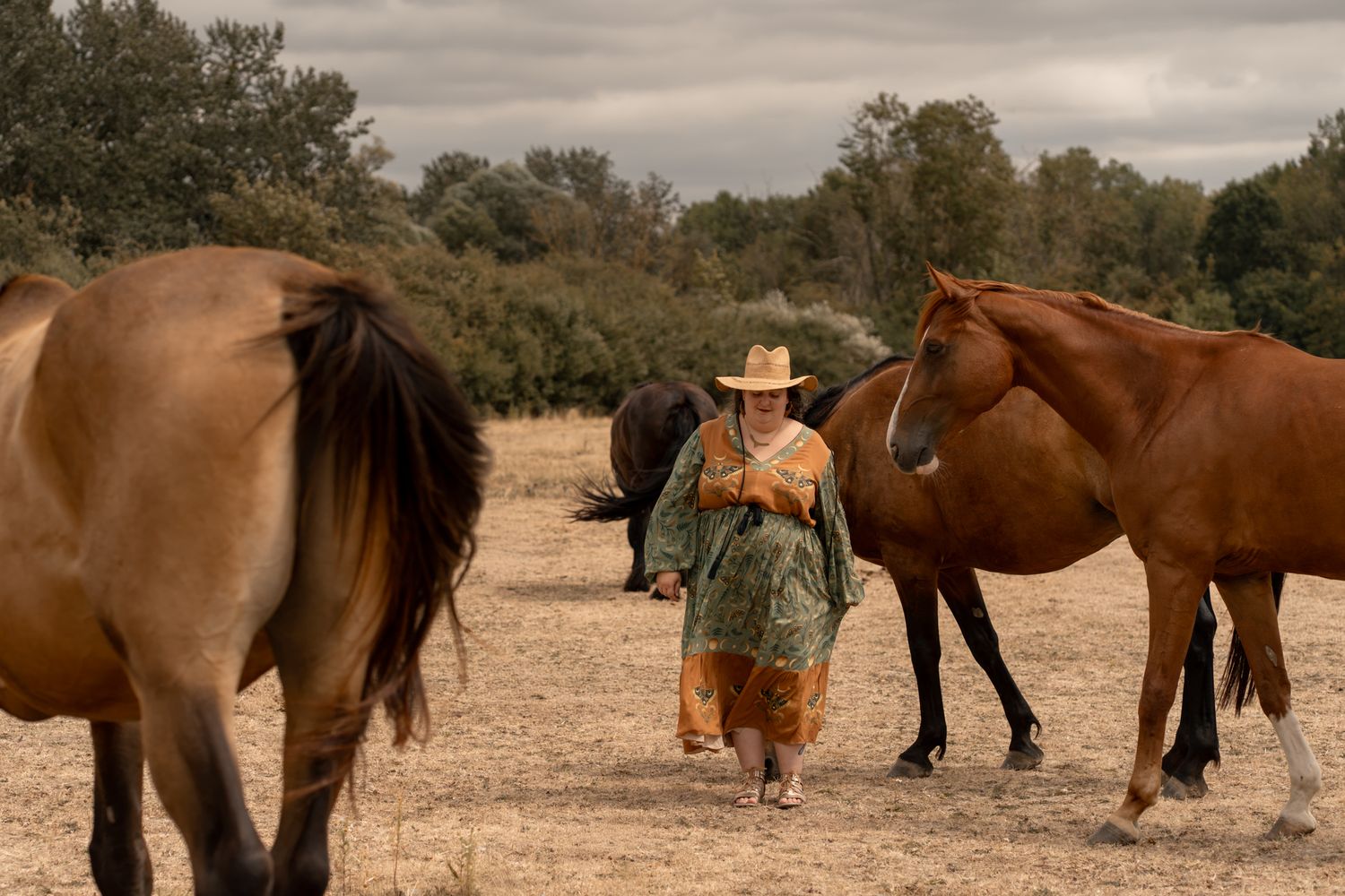 A woman in a straw hat and green outfit walks among brown horses in an open field under cloudy skies.