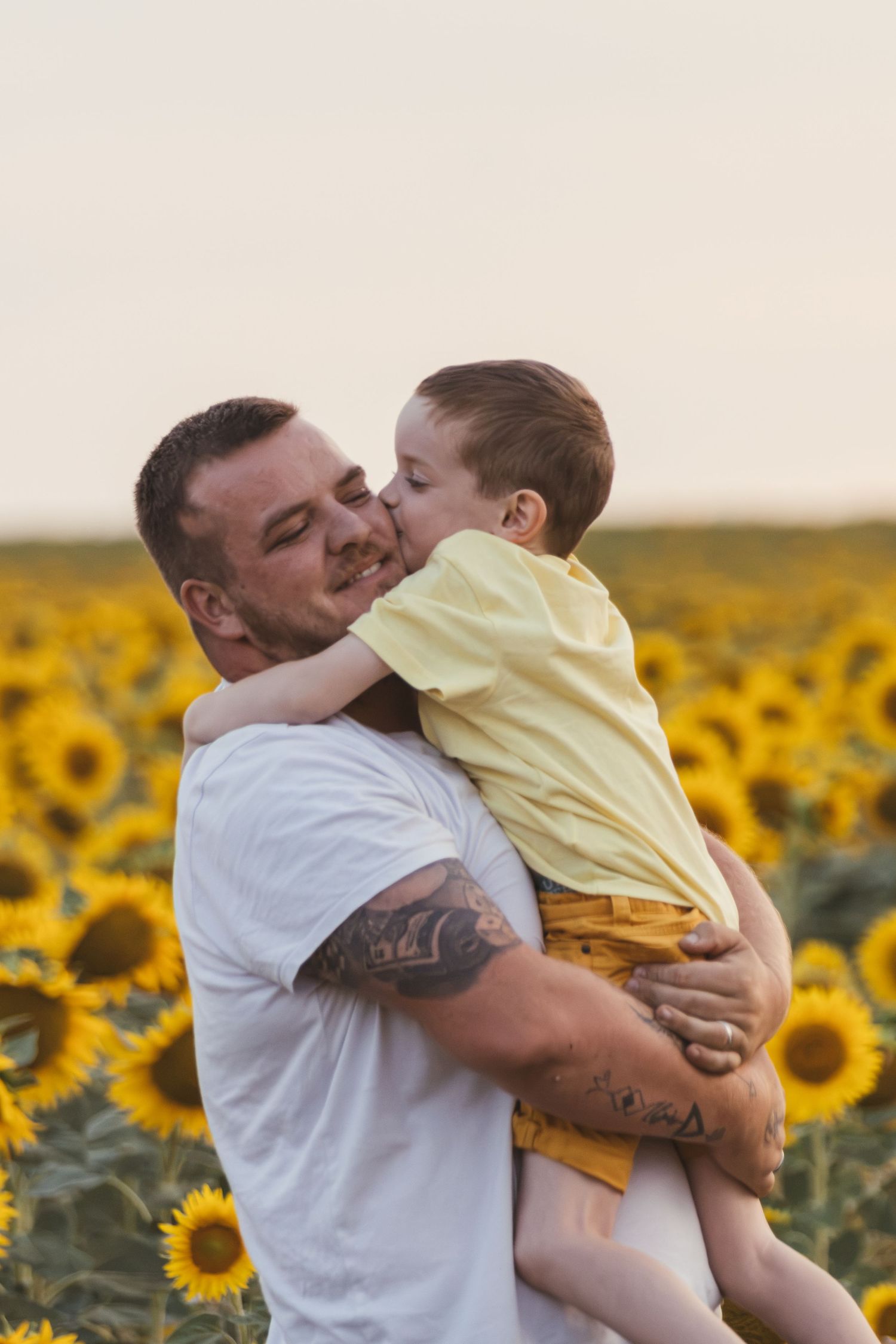 A tattooed man holds a young boy who kisses his cheek in a vibrant sunflower field at sunset.