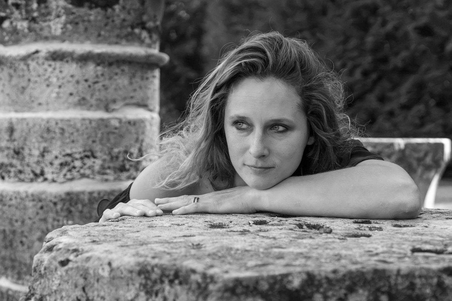Black and white portrait of a woman resting her chin on arms atop a stone wall outdoors.
