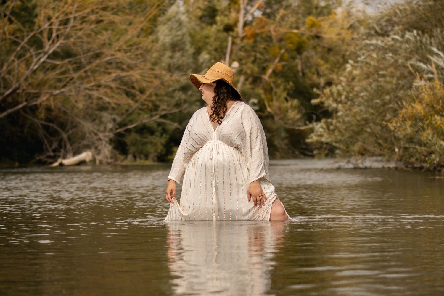 Pregnant woman in white dress and hat standing in river surrounded by trees.