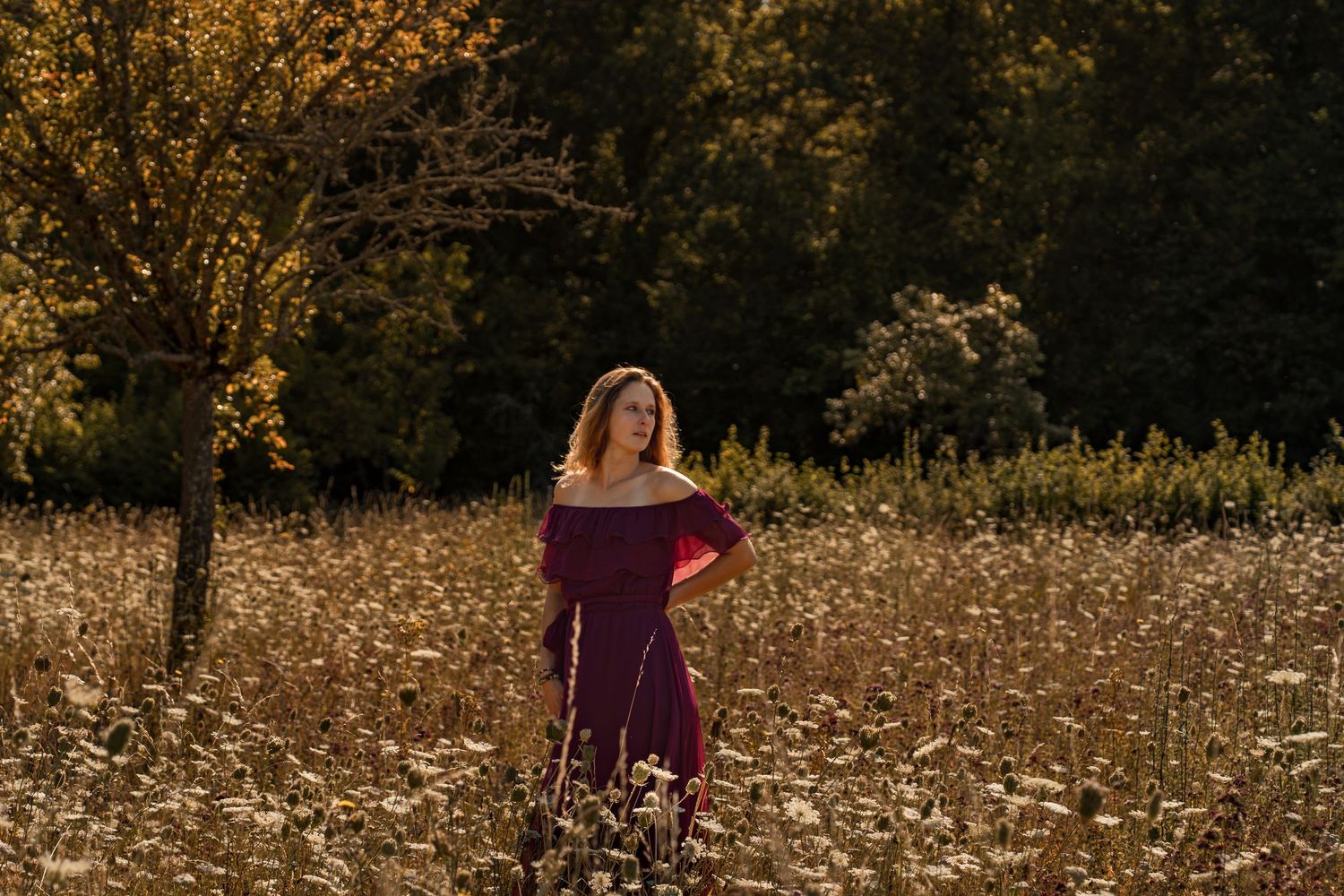 Woman in burgundy off-shoulder dress standing in golden autumn field with wildflowers and colorful trees.