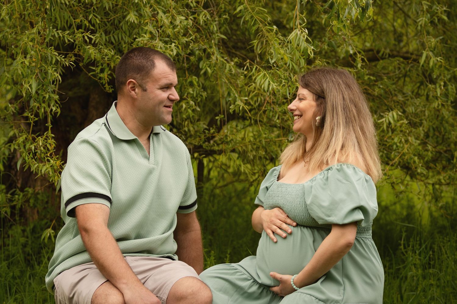 Pregnant couple in matching mint green outfits sitting outdoors under willow tree, sharing a joyful moment.