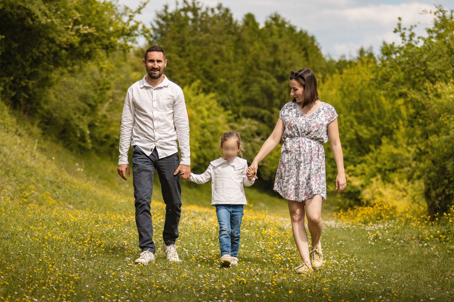 A happy family of three walks hand-in-hand through a lush green meadow with yellow wildflowers on a sunny day.