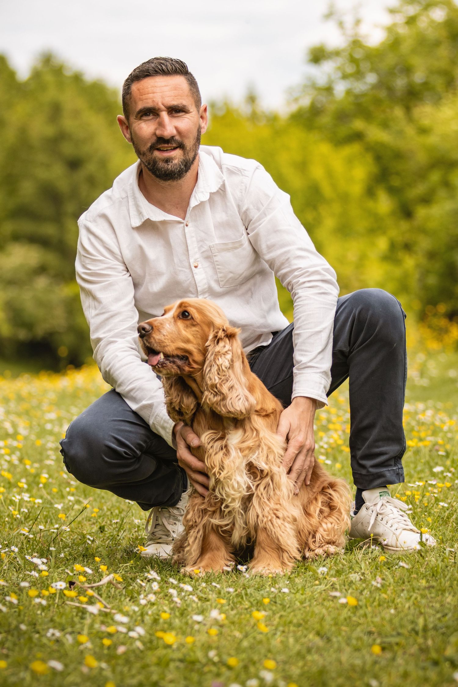 Man in white shirt crouching with a golden Cocker Spaniel in a flower-filled park.