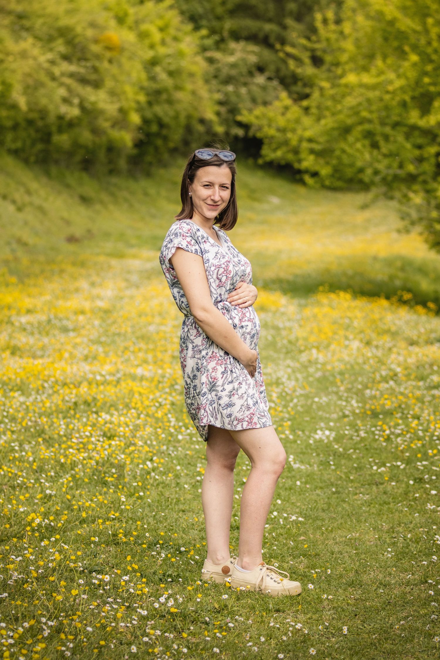 Smiling pregnant woman in floral dress cradling baby bump in sunny wildflower meadow with lush green trees.