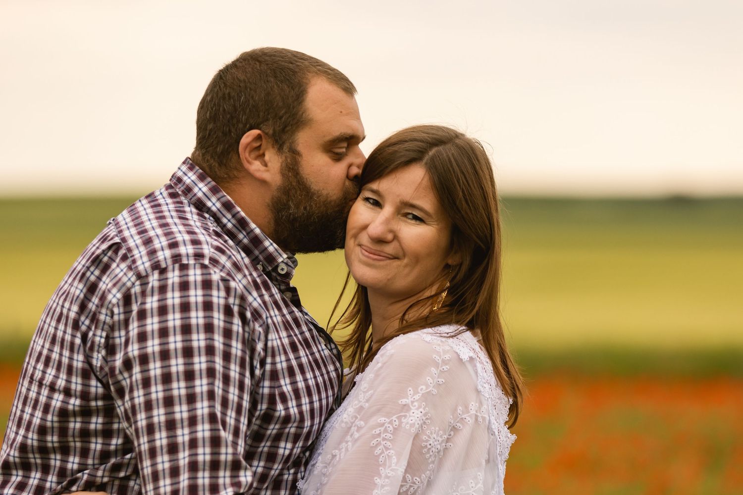 Bearded man kissing woman on the cheek as she smiles in a romantic outdoor field setting.