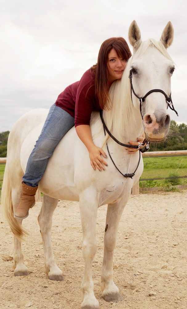 A woman in a red sweater and jeans leans affectionately on a white horse wearing a bridle in an outdoor arena.
