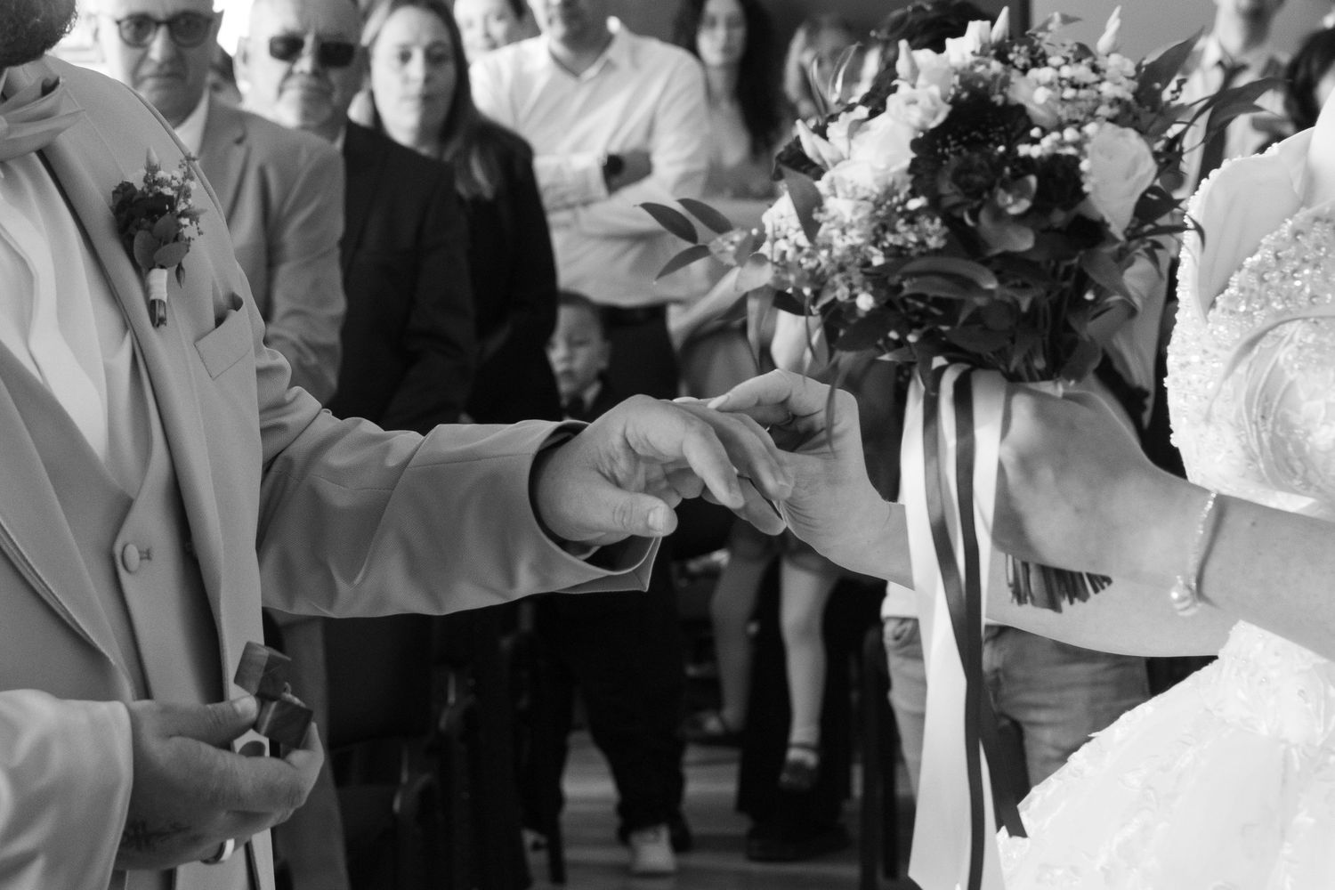 Black and white wedding photo of bride and groom exchanging rings with guests watching in background.