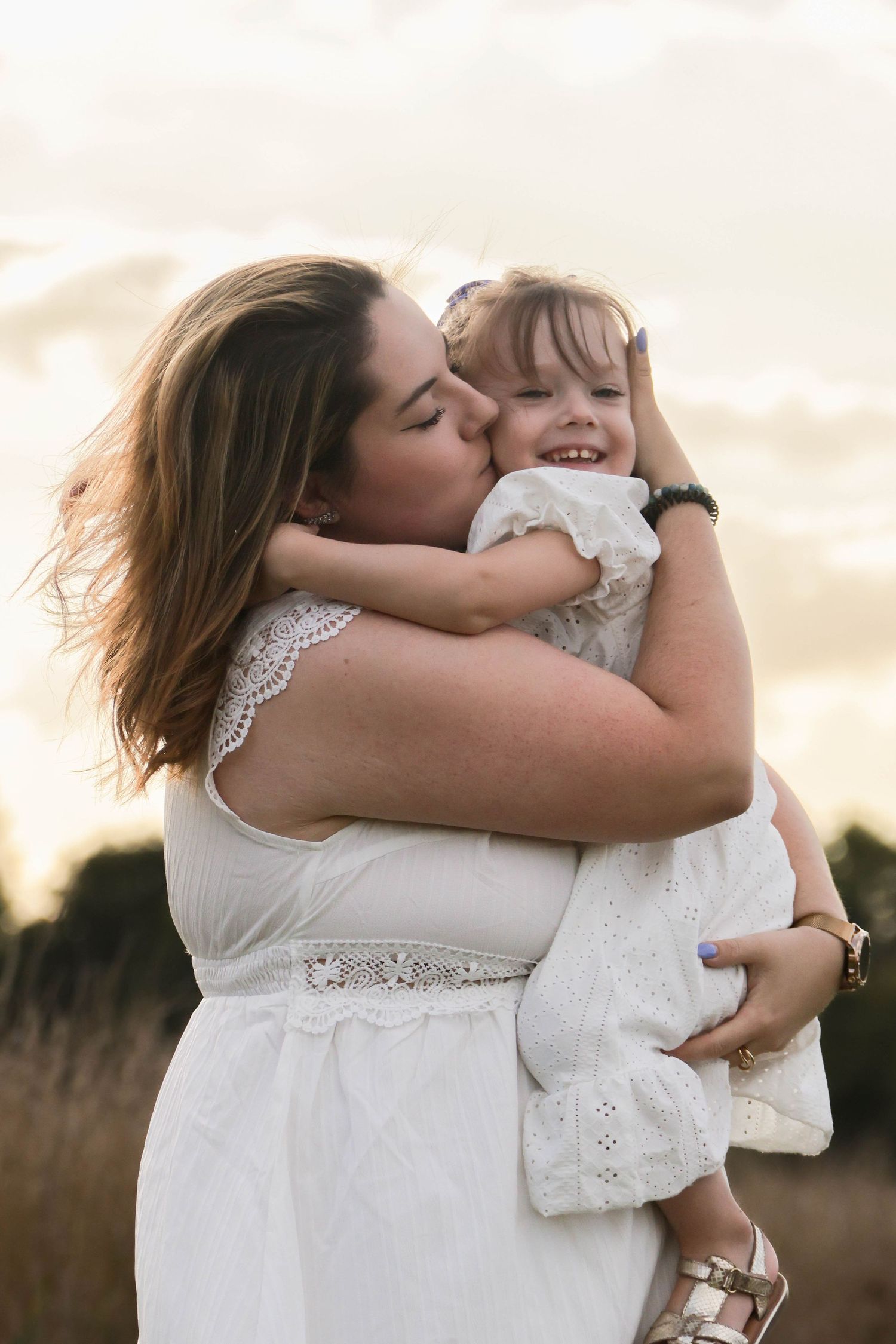 Séance famille lifestyle — maman en robe blanche embrassant tendrement sa petite fille souriante au coucher du soleil, photographe rethel