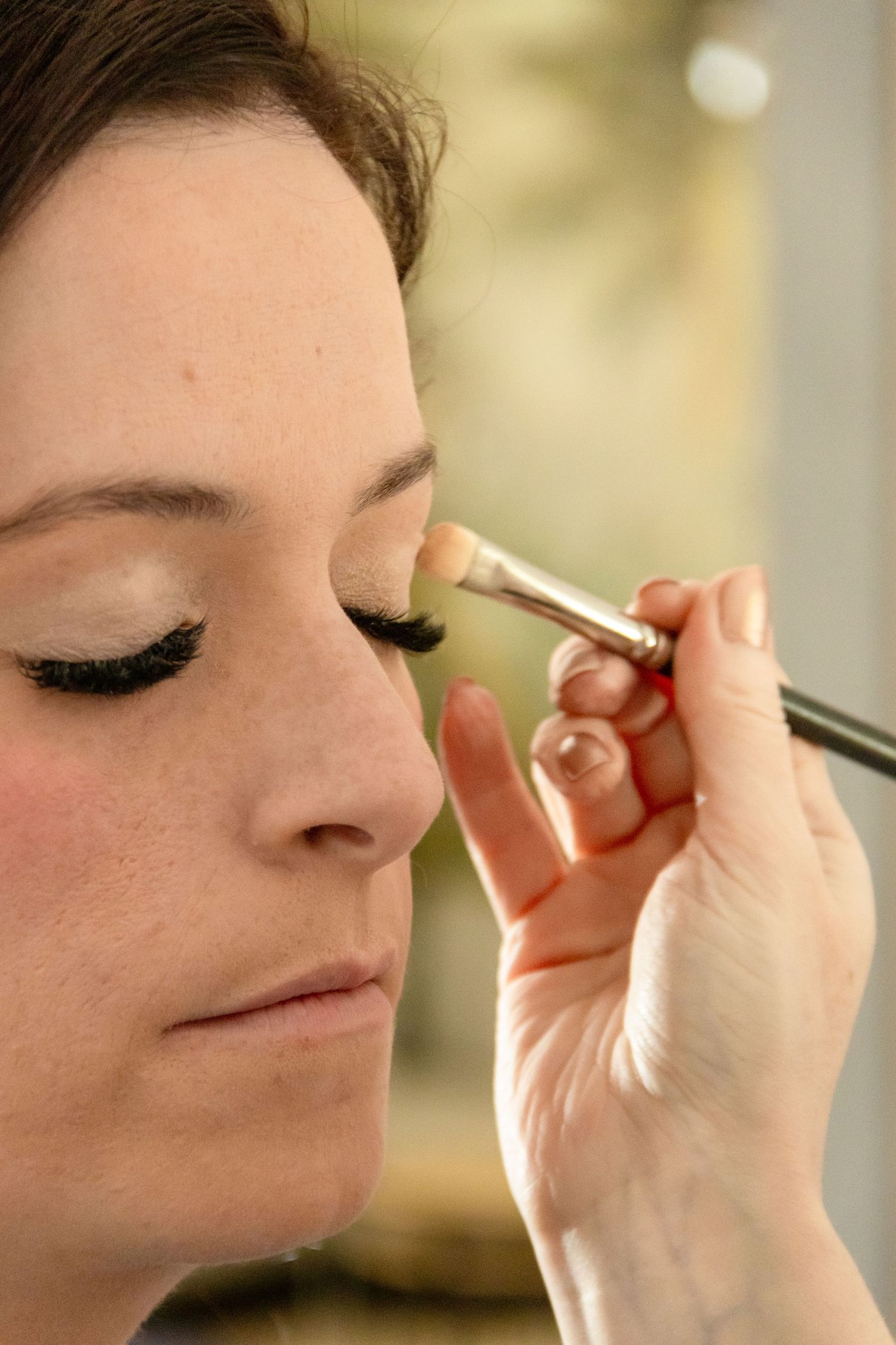 A makeup artist applying eyeshadow with a brush to a woman with false lashes and closed eyes.
