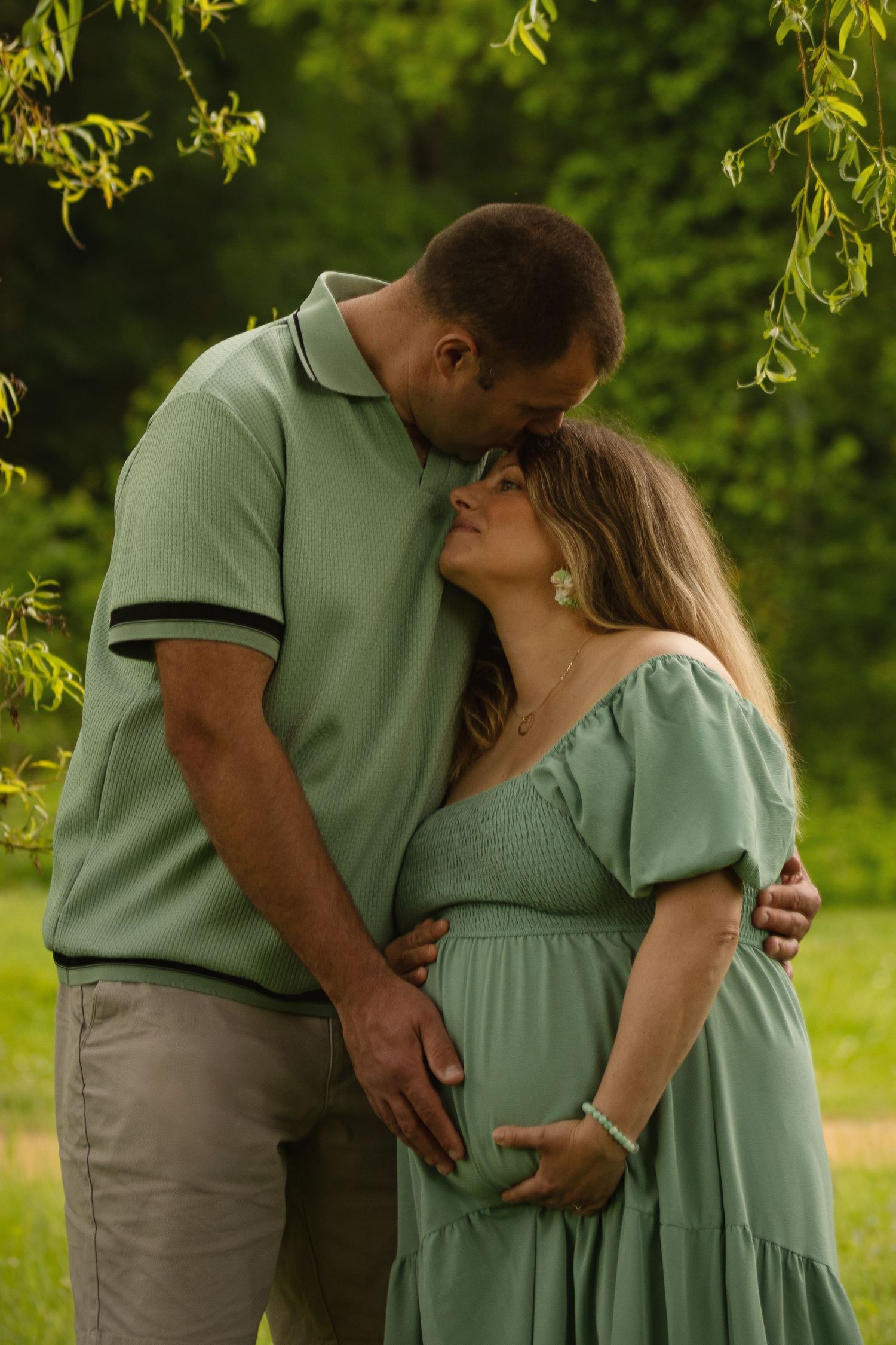 A couple in matching green outfits share a tender moment outdoors, with the man kissing a pregnant woman's forehead.