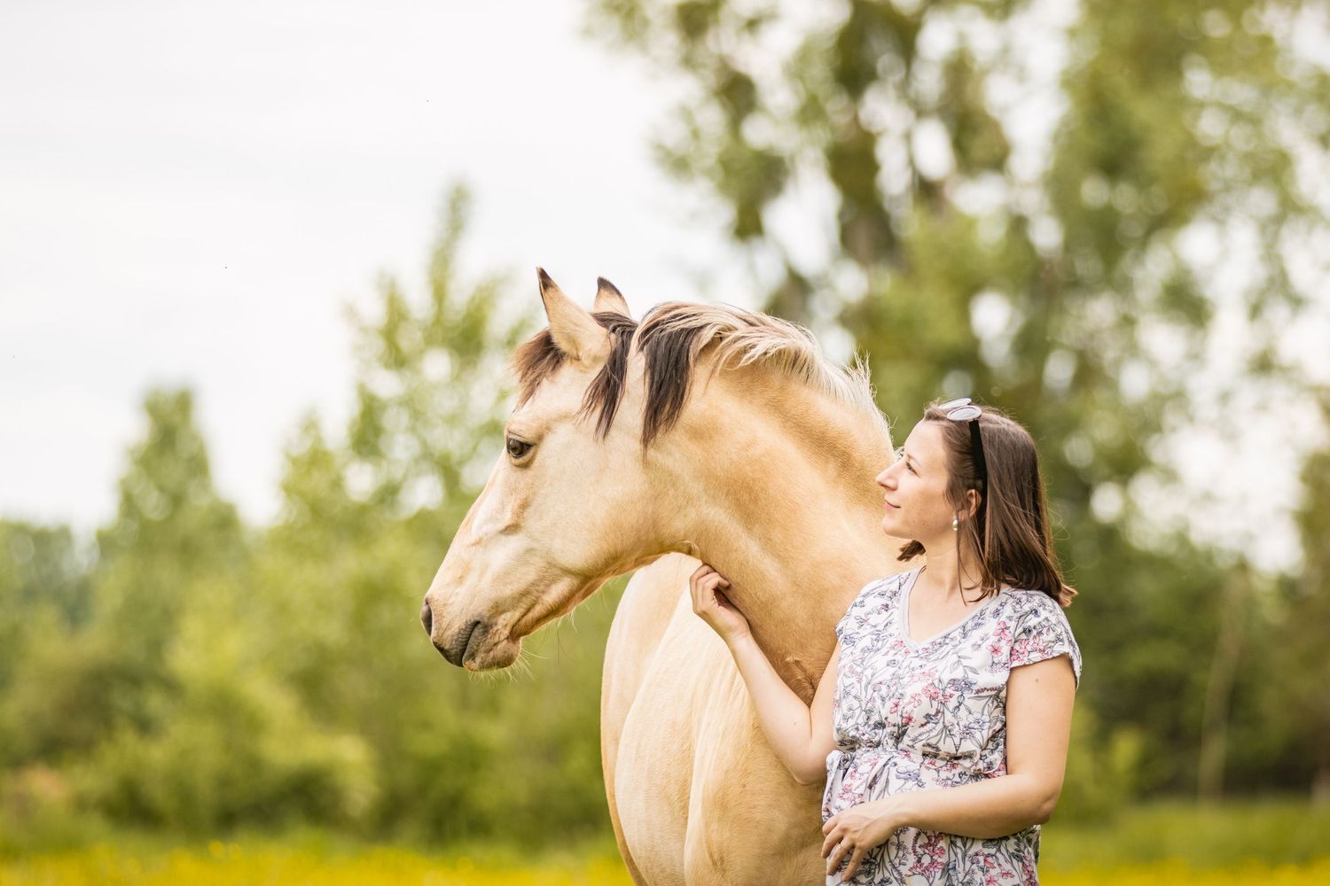 A young woman smiles affectionately at a palomino horse in a lush green field.