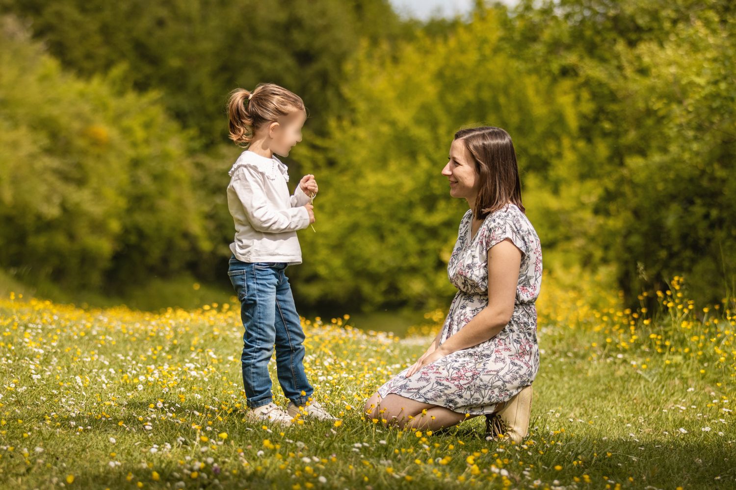 A young girl offers a flower to a woman kneeling in a sunny meadow with yellow wildflowers and green trees.
