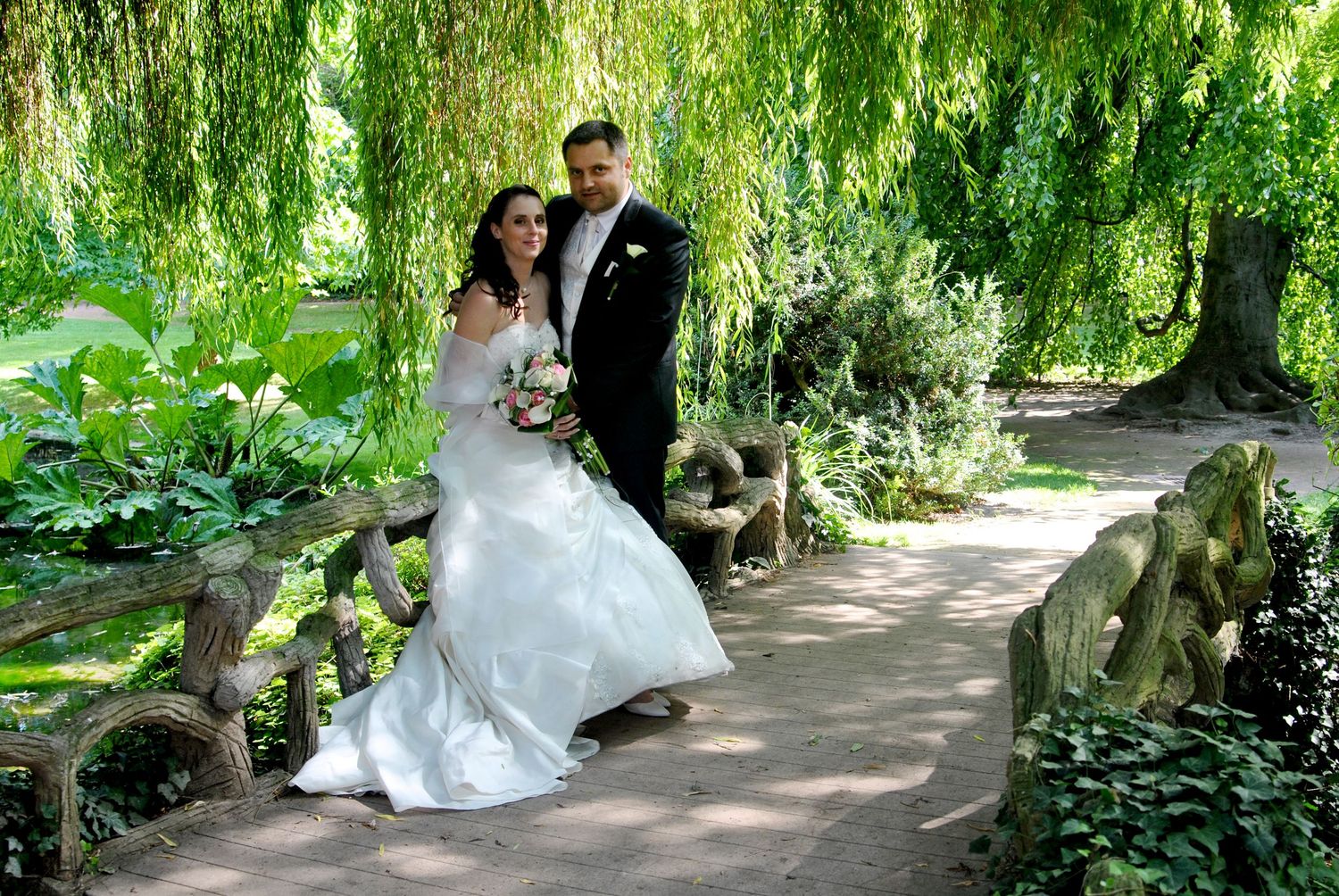 Bride and groom pose romantically on a garden bridge under green trees.