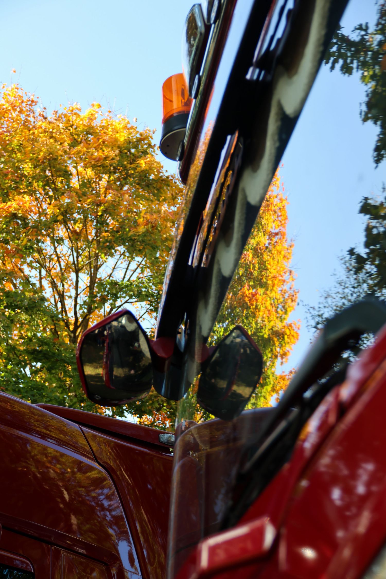 Low angle view of a red truck cab with side mirrors and amber warning light against autumn trees and blue sky.
