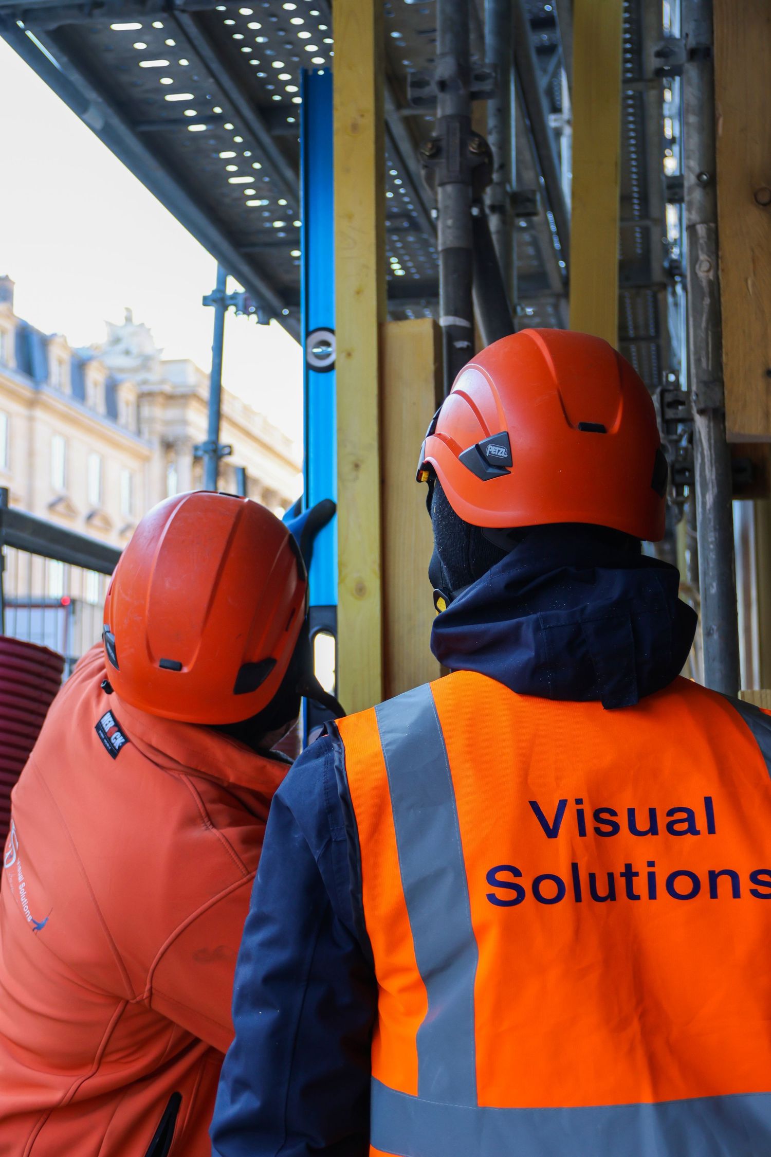Two construction workers in orange helmets and safety vests with Visual Solutions logo inspect a building structure.