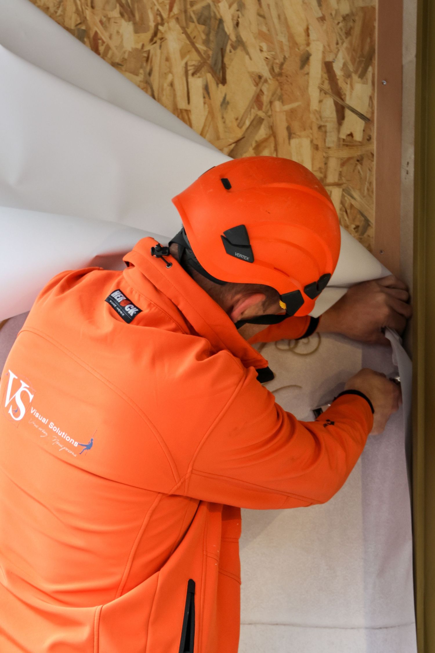 Worker in orange safety gear and helmet installs insulation material against an OSB board wall.