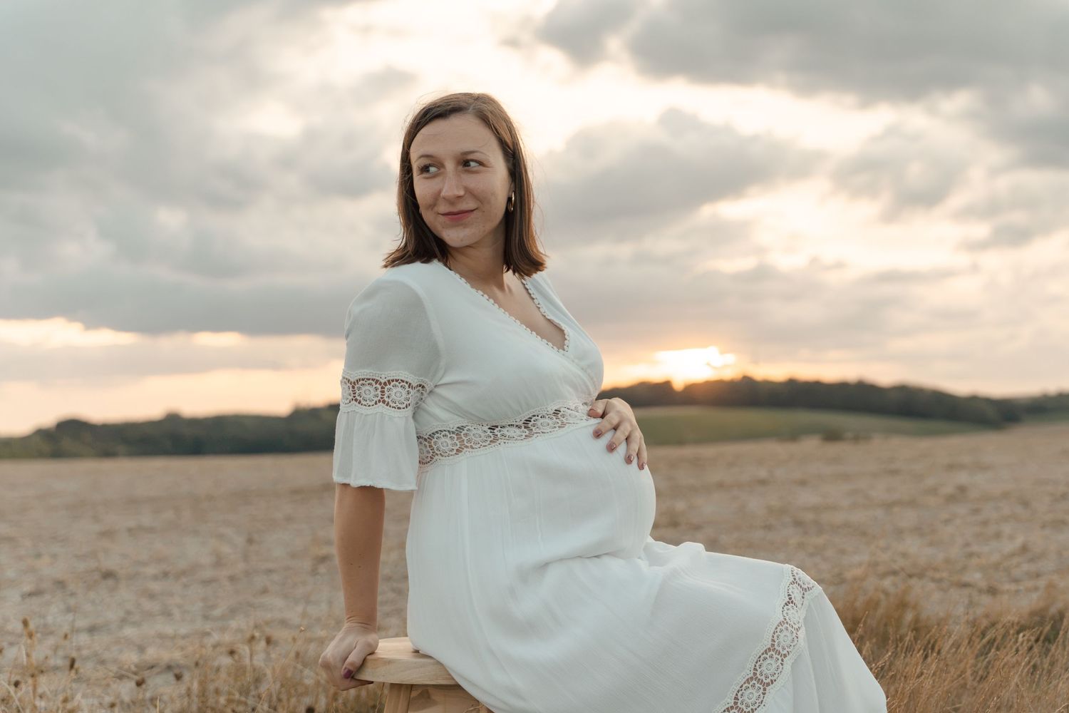 Pregnant woman in white dress sitting in golden field at sunset, cradling baby bump.