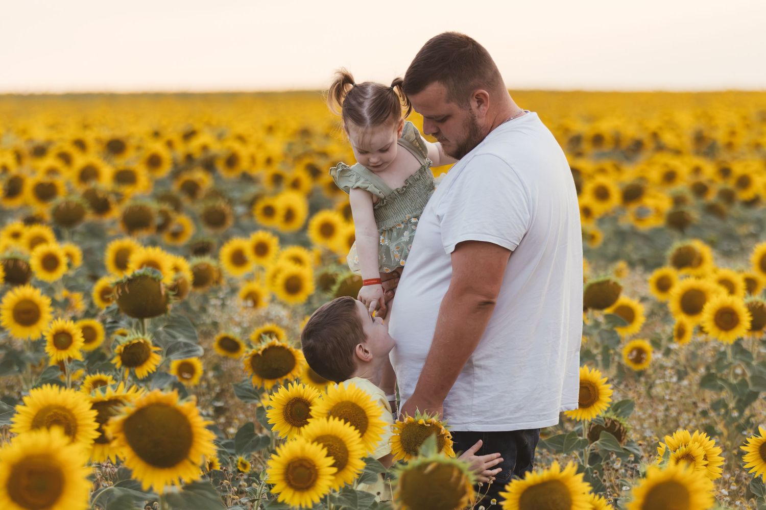 Father holding young daughter in a vast sunflower field at sunset, with son hugging his waist.