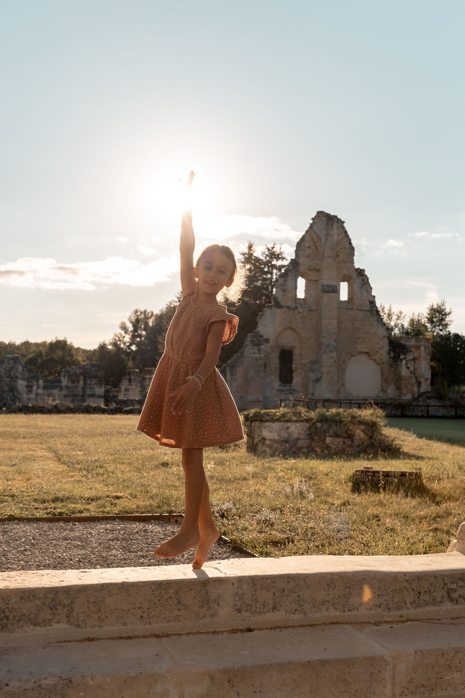 Young girl in pink dress poses ballet-style on stone ledge with sunlit ruins in background.