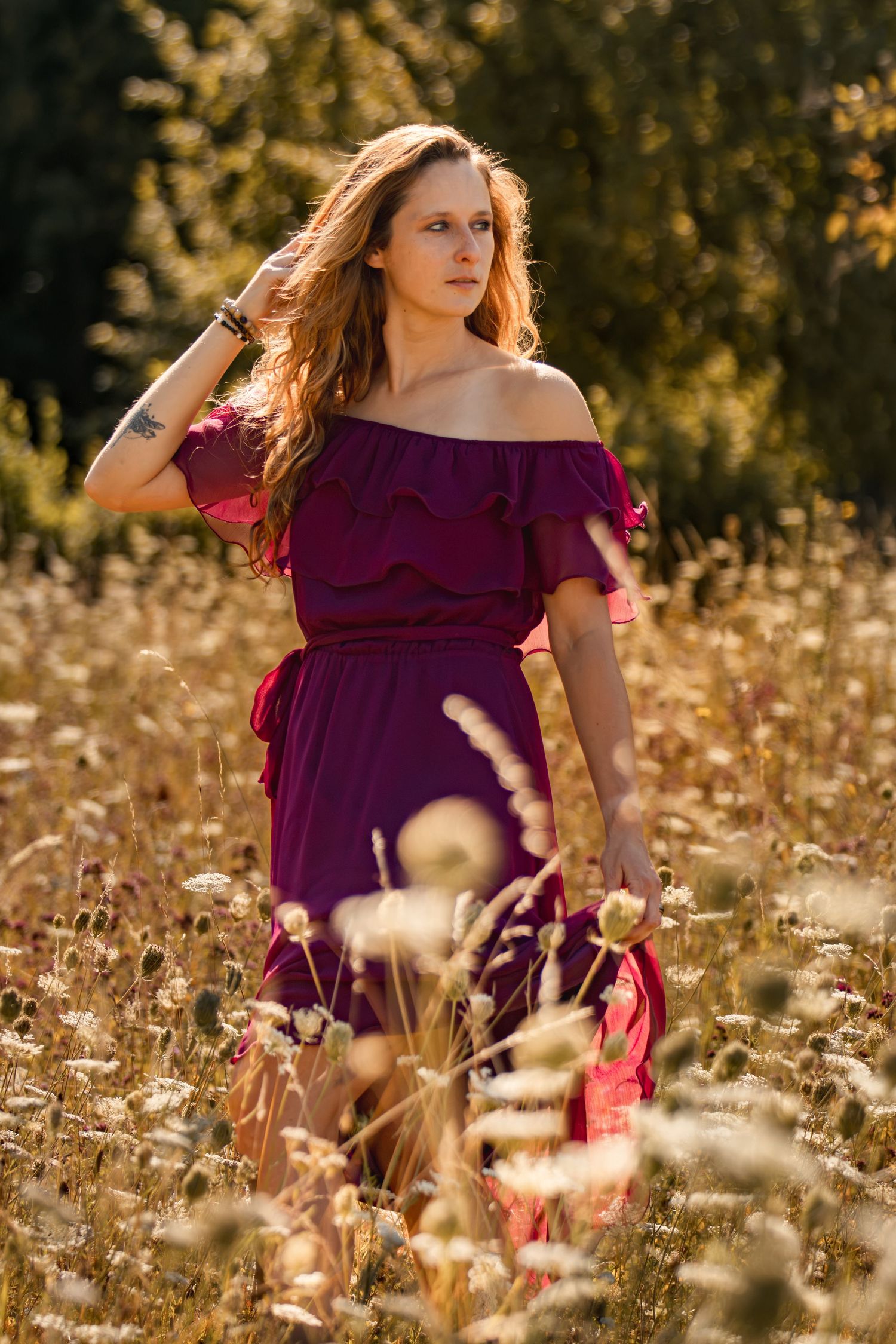 Woman in burgundy off-shoulder dress standing in a sunny wildflower field with long auburn hair.