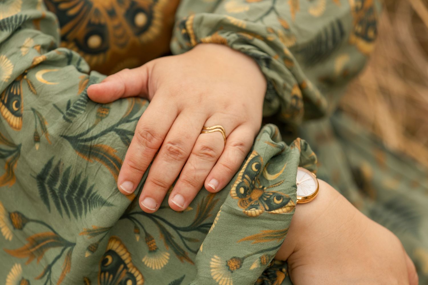 Close-up of a baby hand resting on an adult's green floral dress, with gold rings and bracelet accessories.