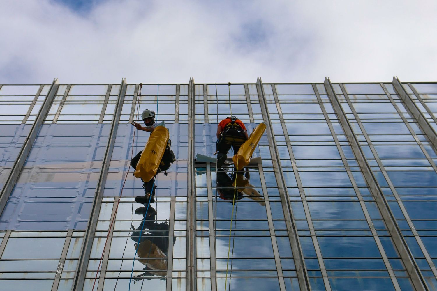 Two window washers rappelling on a glass skyscraper facade, cleaning windows against a cloudy blue sky.