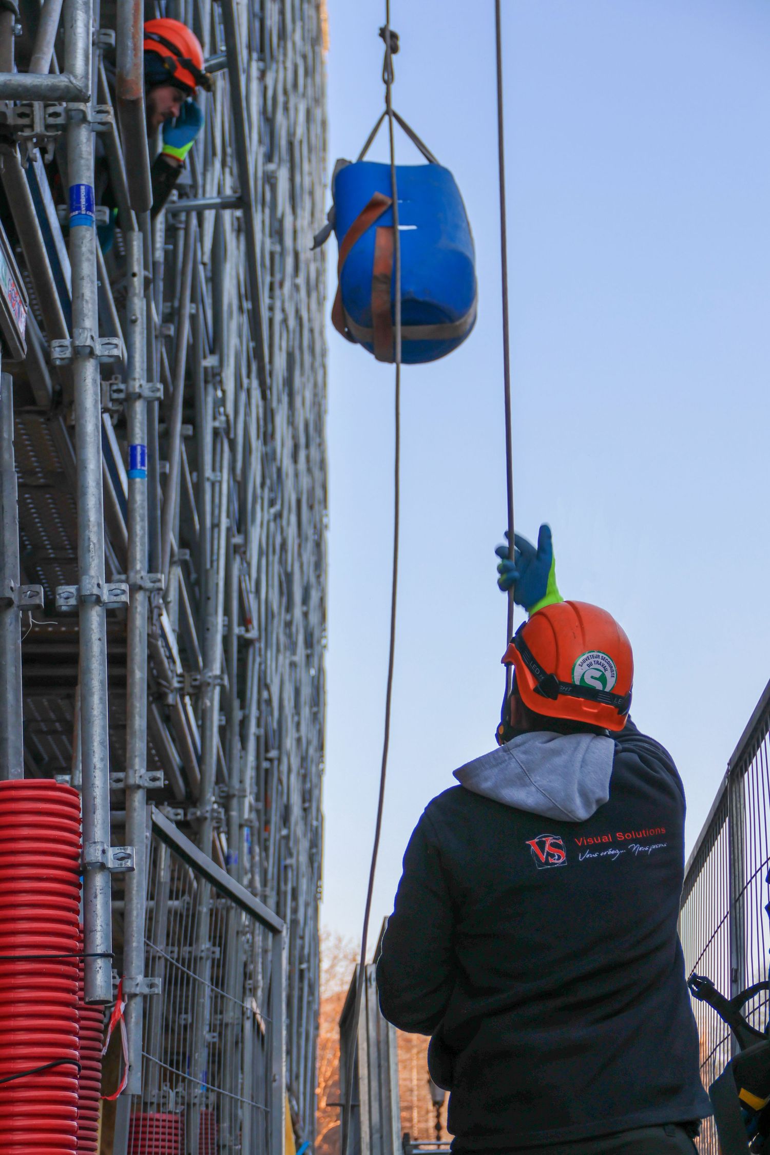 Construction workers in orange helmets using a crane to hoist a blue container up scaffolding on a building site.