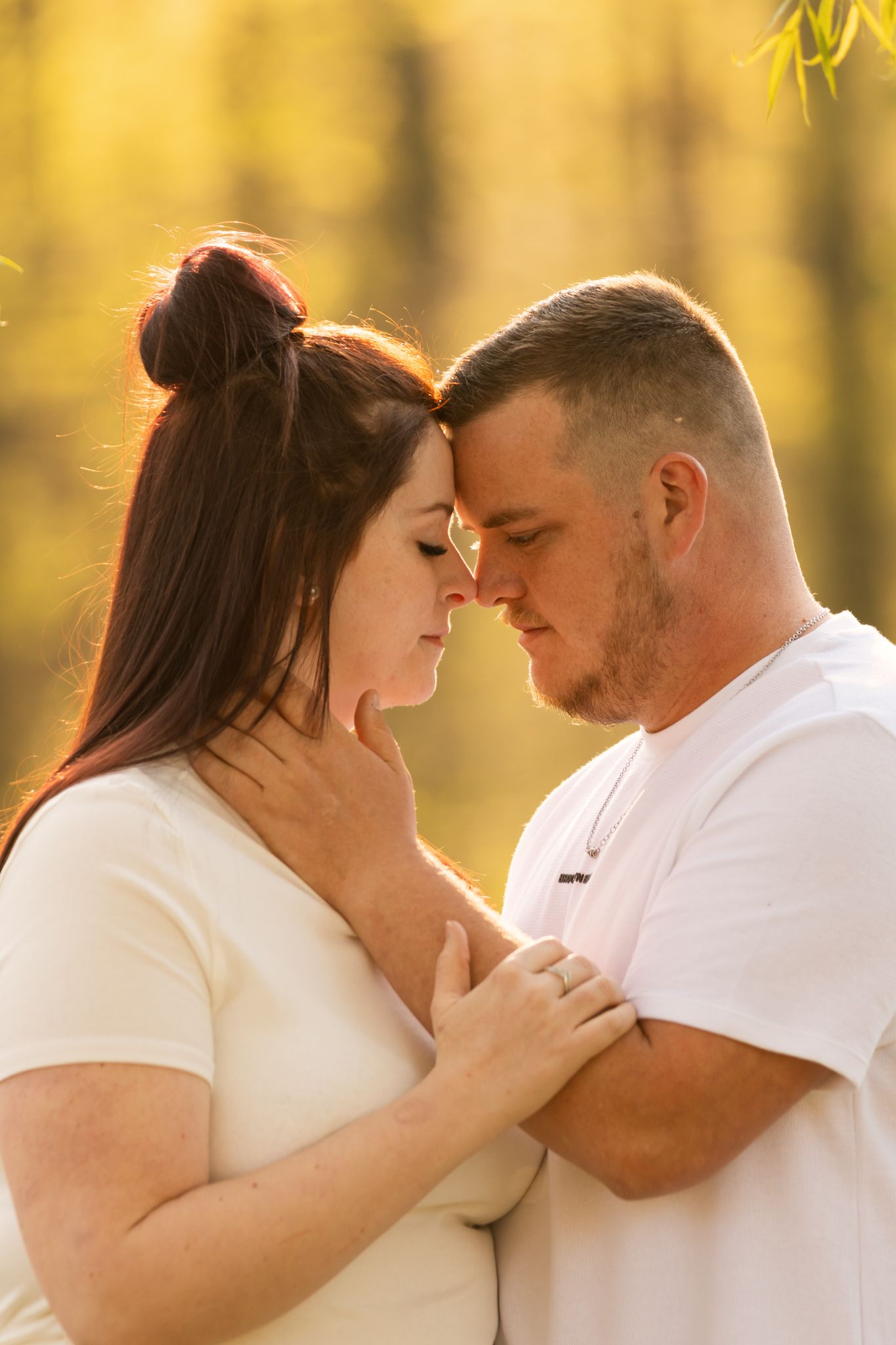 A couple shares an intimate forehead-to-forehead moment outdoors, bathed in warm golden light among autumn trees.