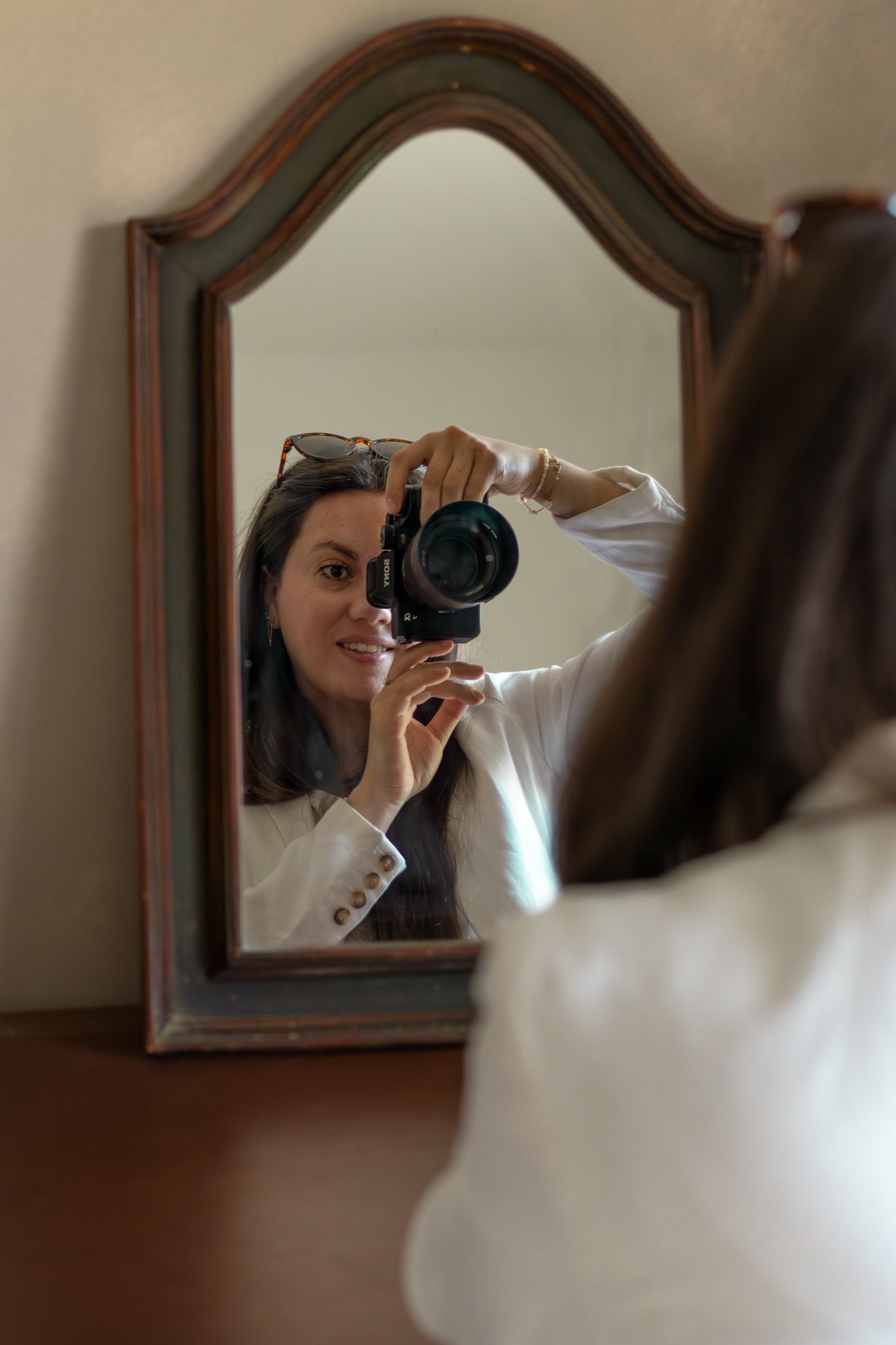 Woman taking a mirror selfie with a DSLR camera, smiling, wearing a white blazer with sunglasses on her head.