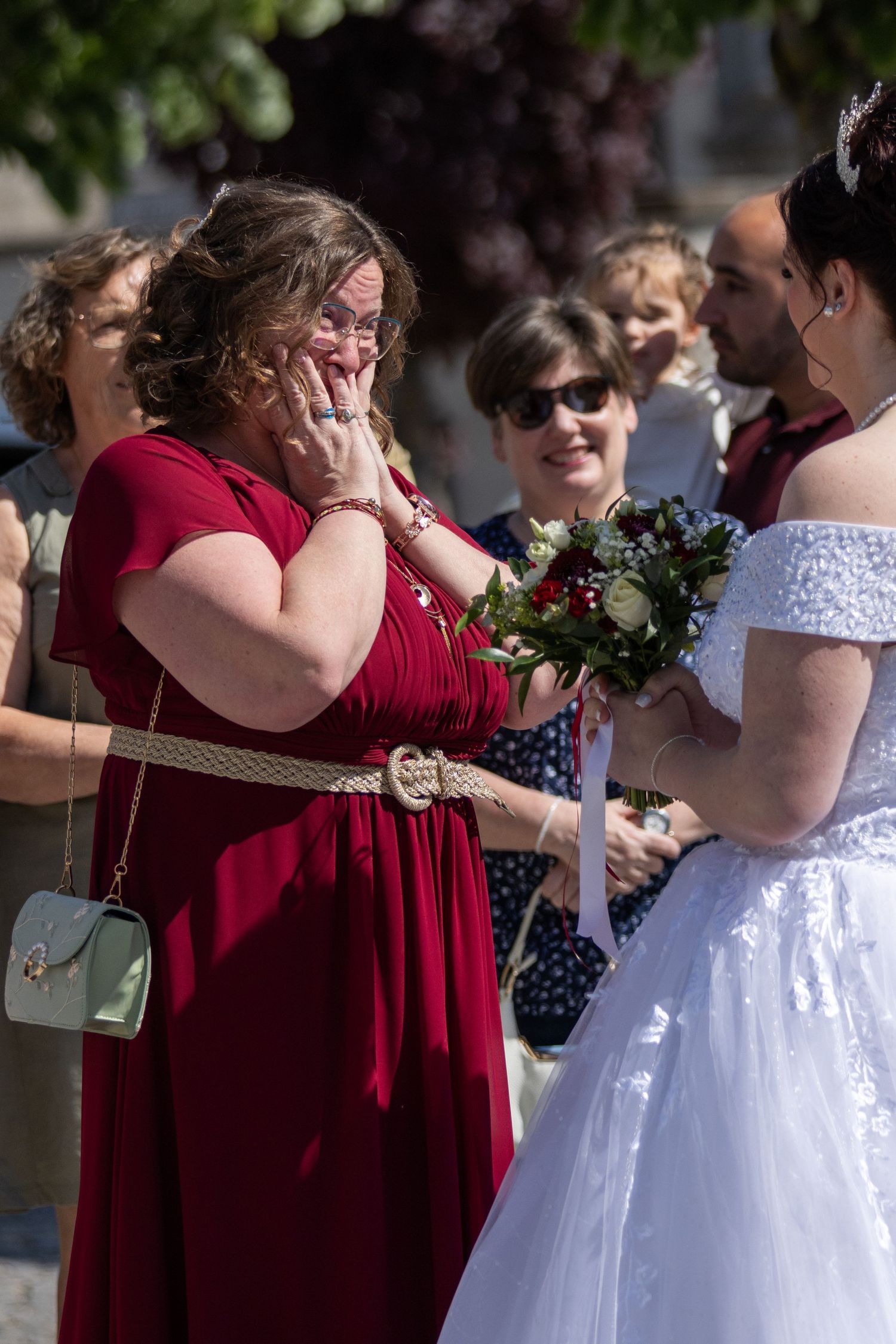 Emotional woman in red dress wiping tears at outdoor wedding ceremony while bride in white gown holds bouquet.