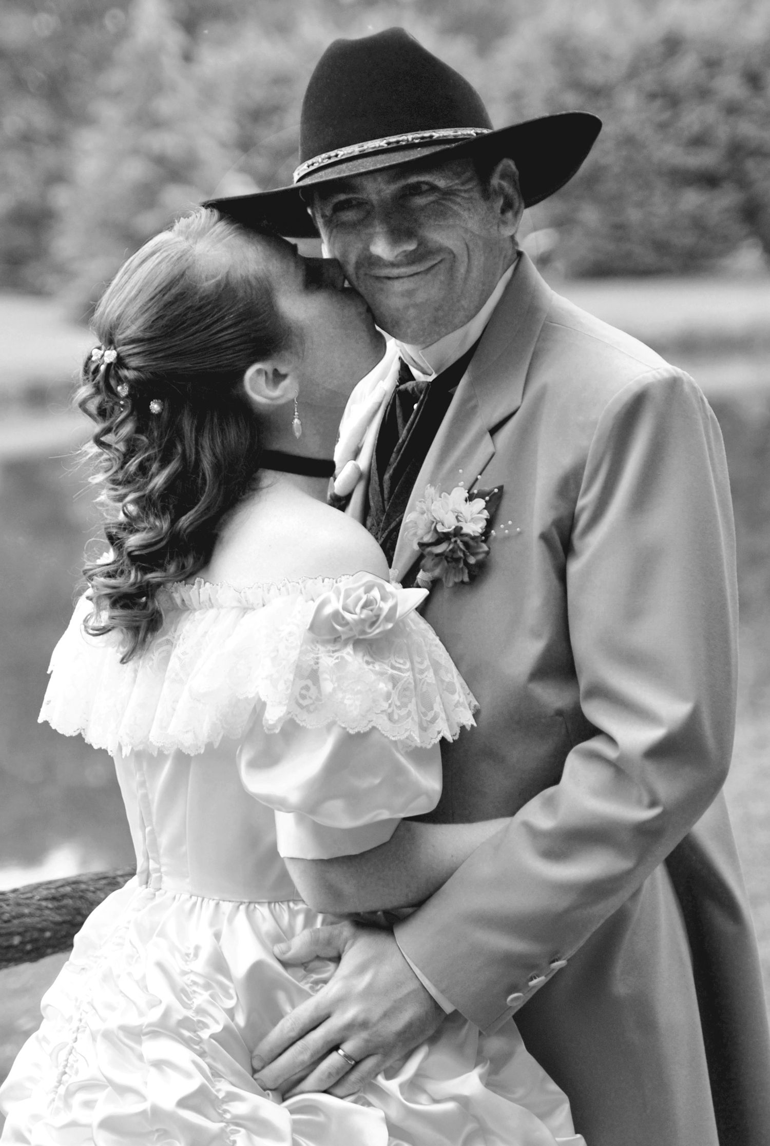 Black and white photo of western bride kissing groom's cheek as he smiles in cowboy hat and tan suit.