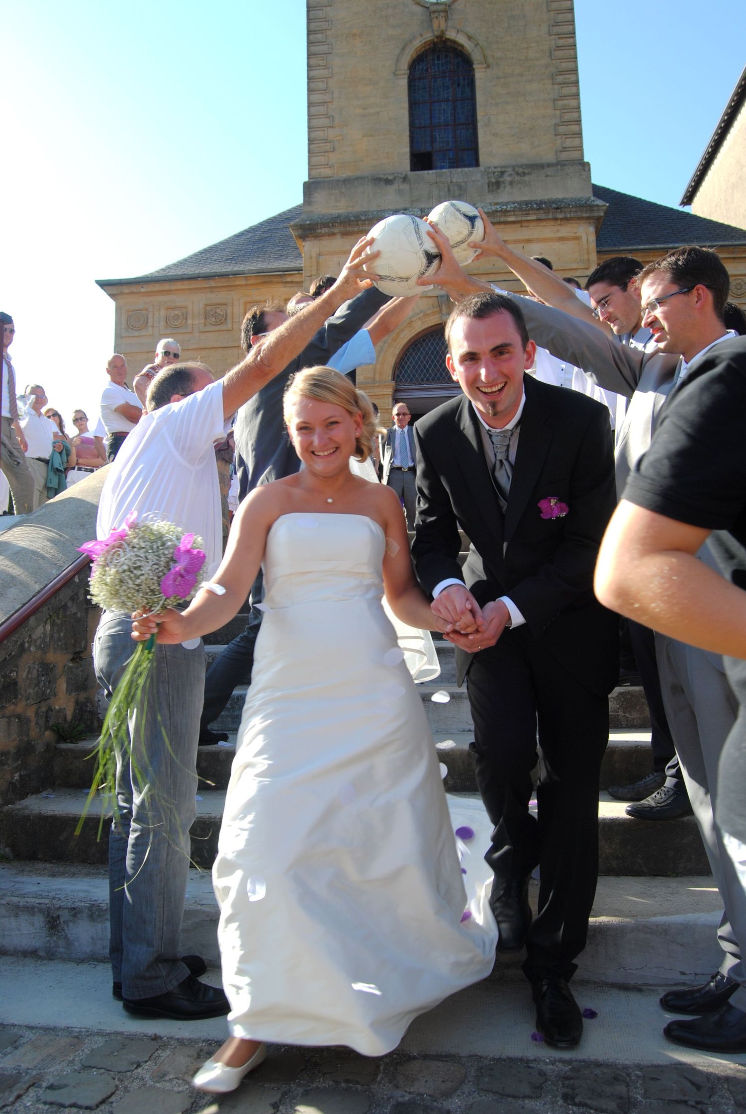 Bride and groom descend church steps as guests hold soccer ball overhead during outdoor wedding ceremony.