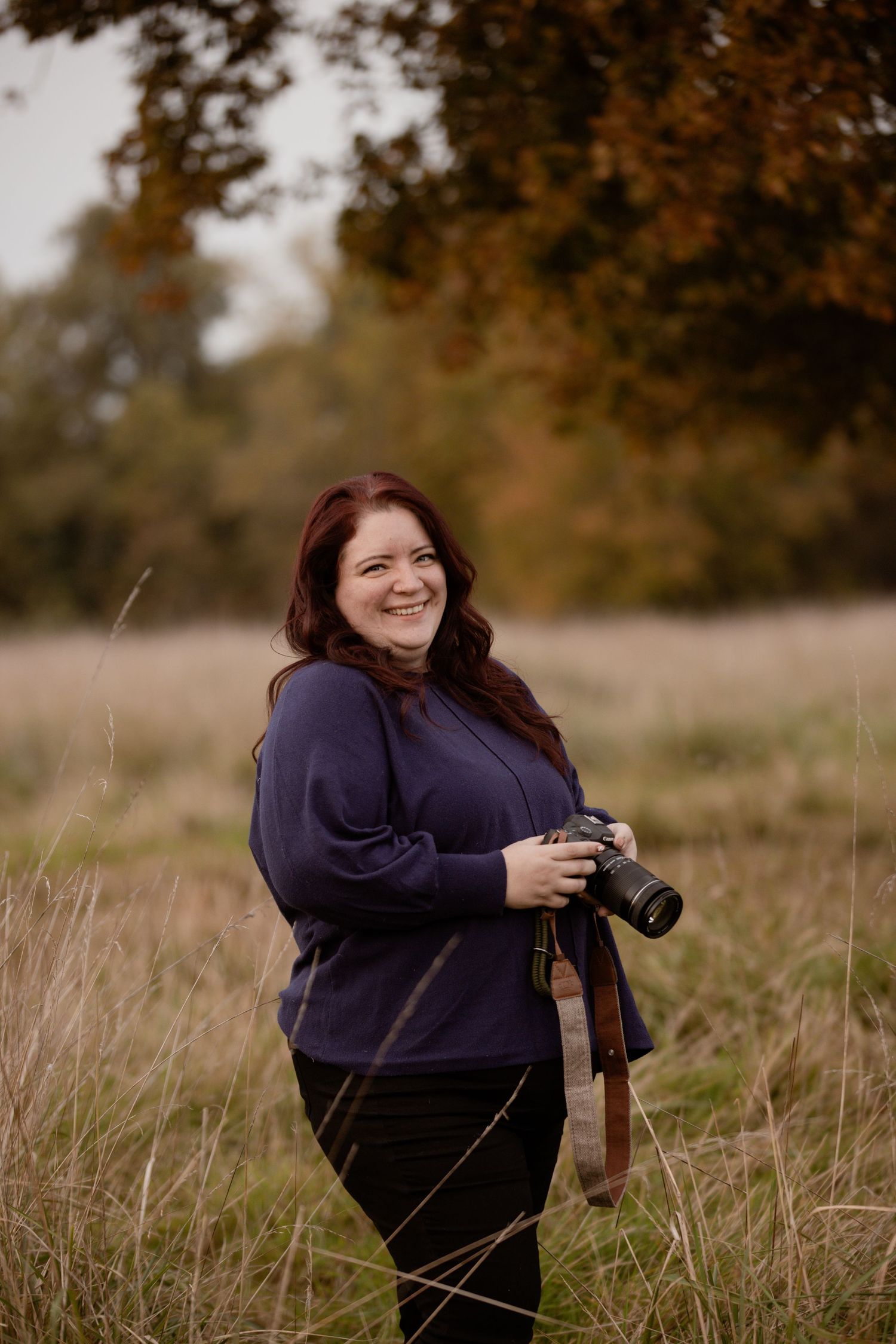 A smiling woman with red hair holds a professional camera while standing in an autumn meadow.