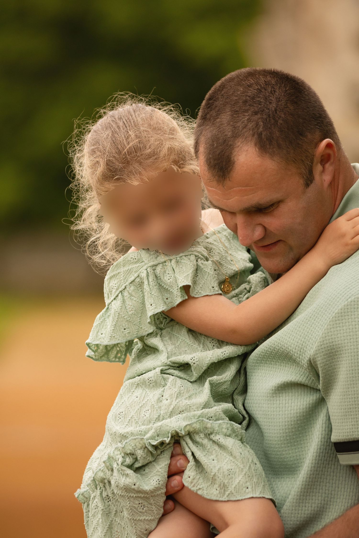 A father tenderly holds his young blonde daughter in matching green outfits outdoors.