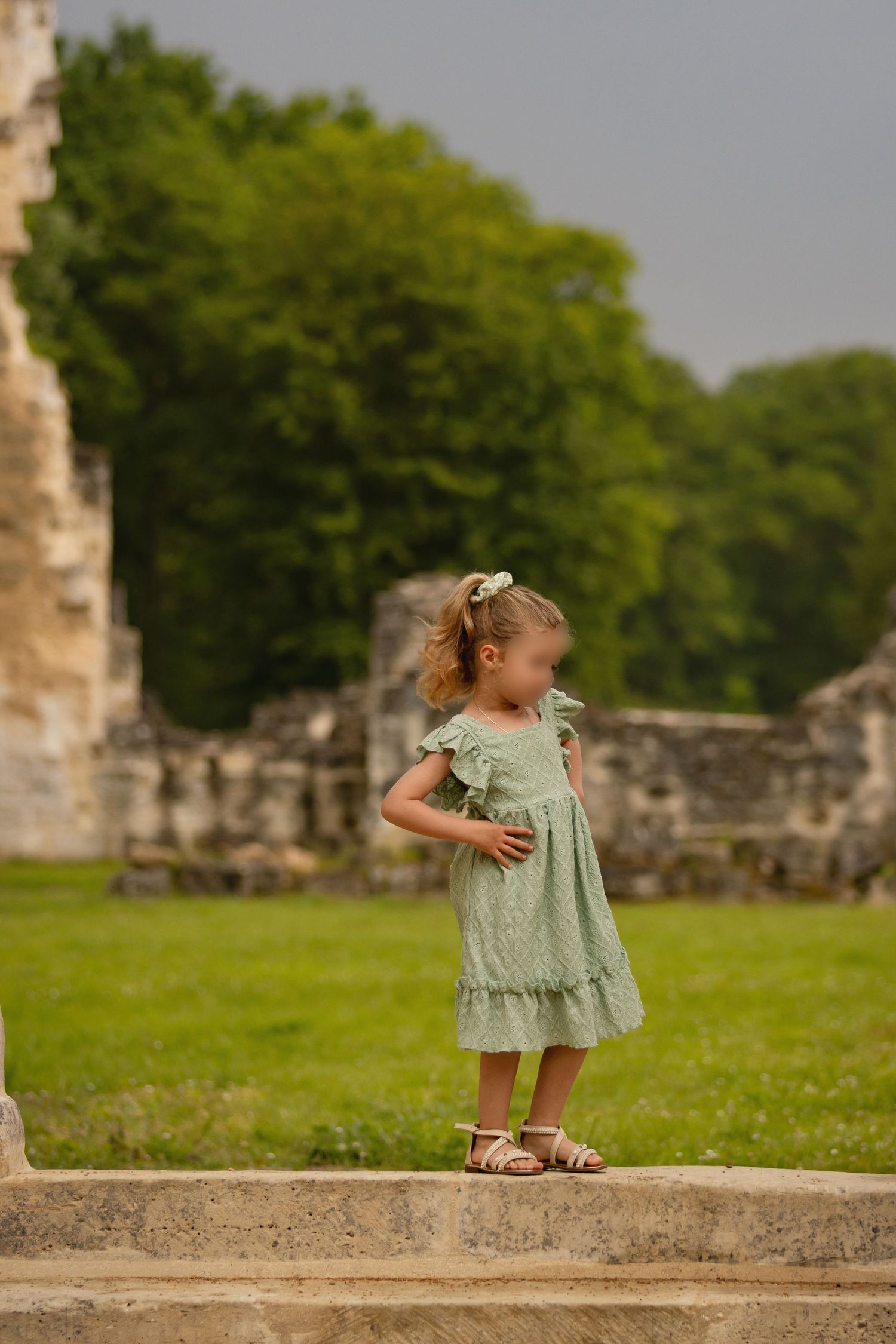 Young girl in green dress and sandals standing on stone ledge near ancient ruins with lush green trees in background.