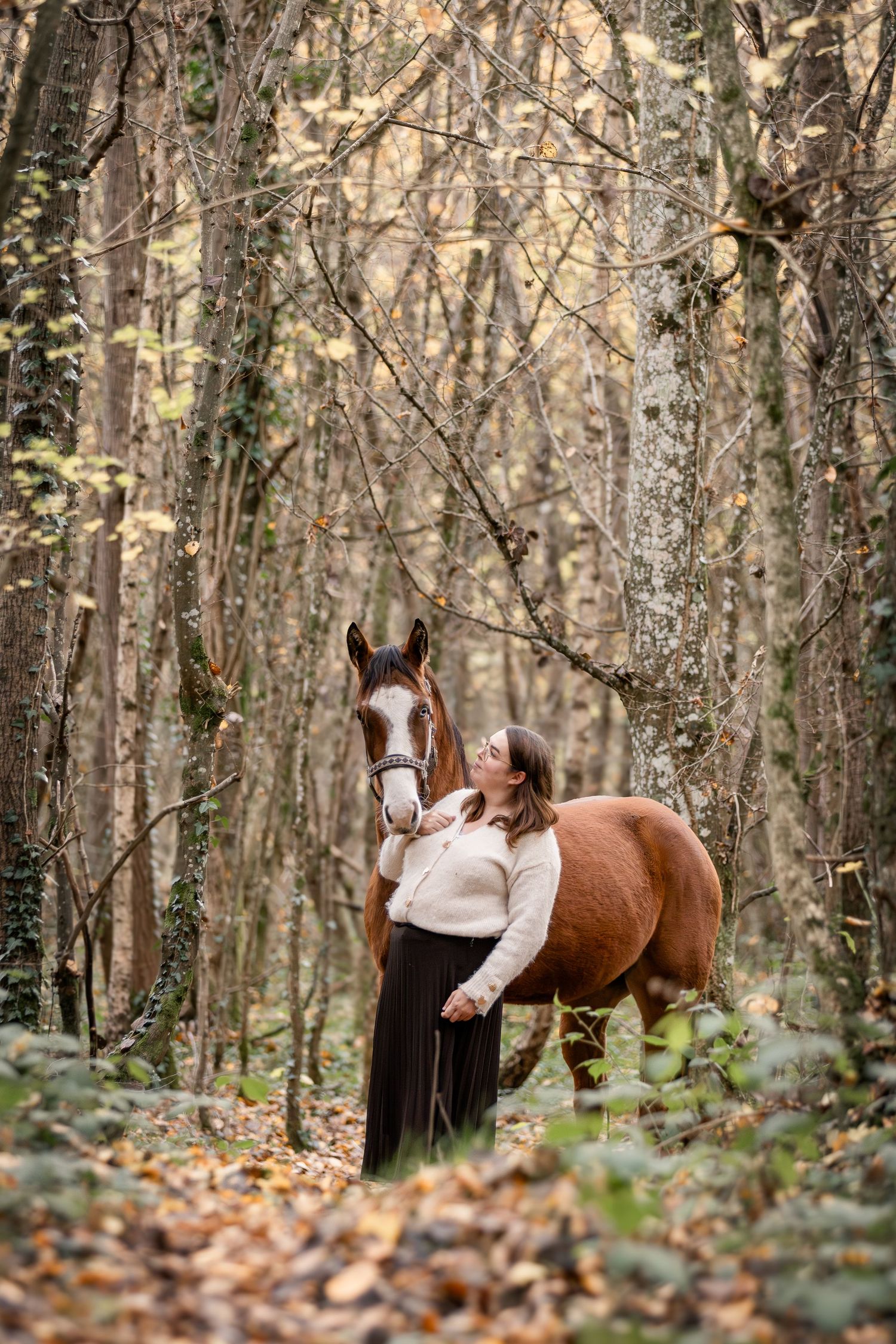 A woman in a white sweater and dark skirt stands close to a chestnut horse in an autumn woodland.