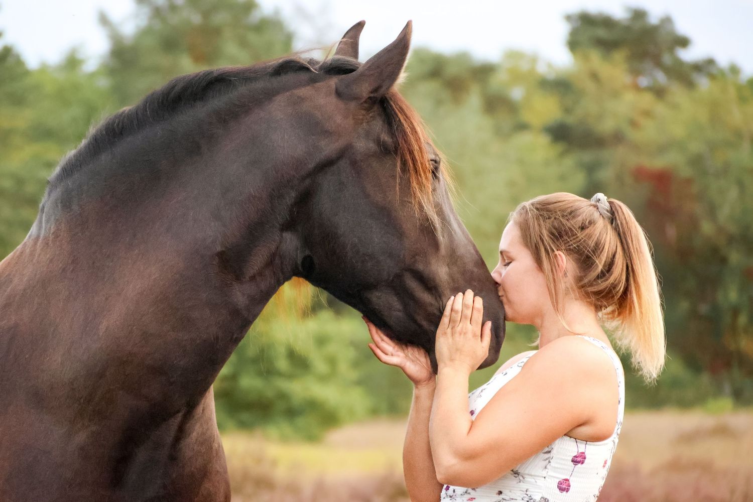 A young blonde woman tenderly touching noses with a dark brown horse in a natural outdoor setting.