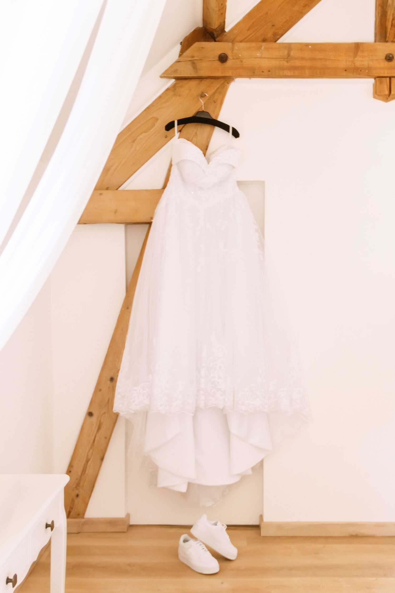 White strapless wedding dress hanging on hanger with white sneakers below, displayed in rustic room with wooden beams.