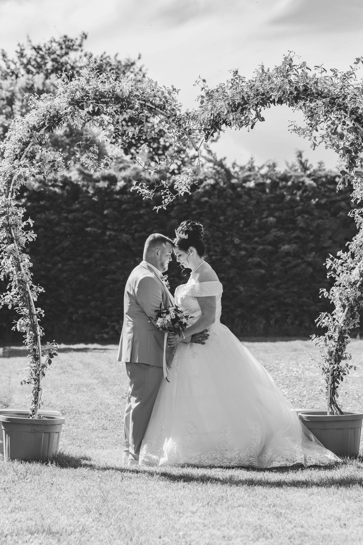 Black and white photo of a bride and groom standing under a floral arch in a garden on their wedding day.