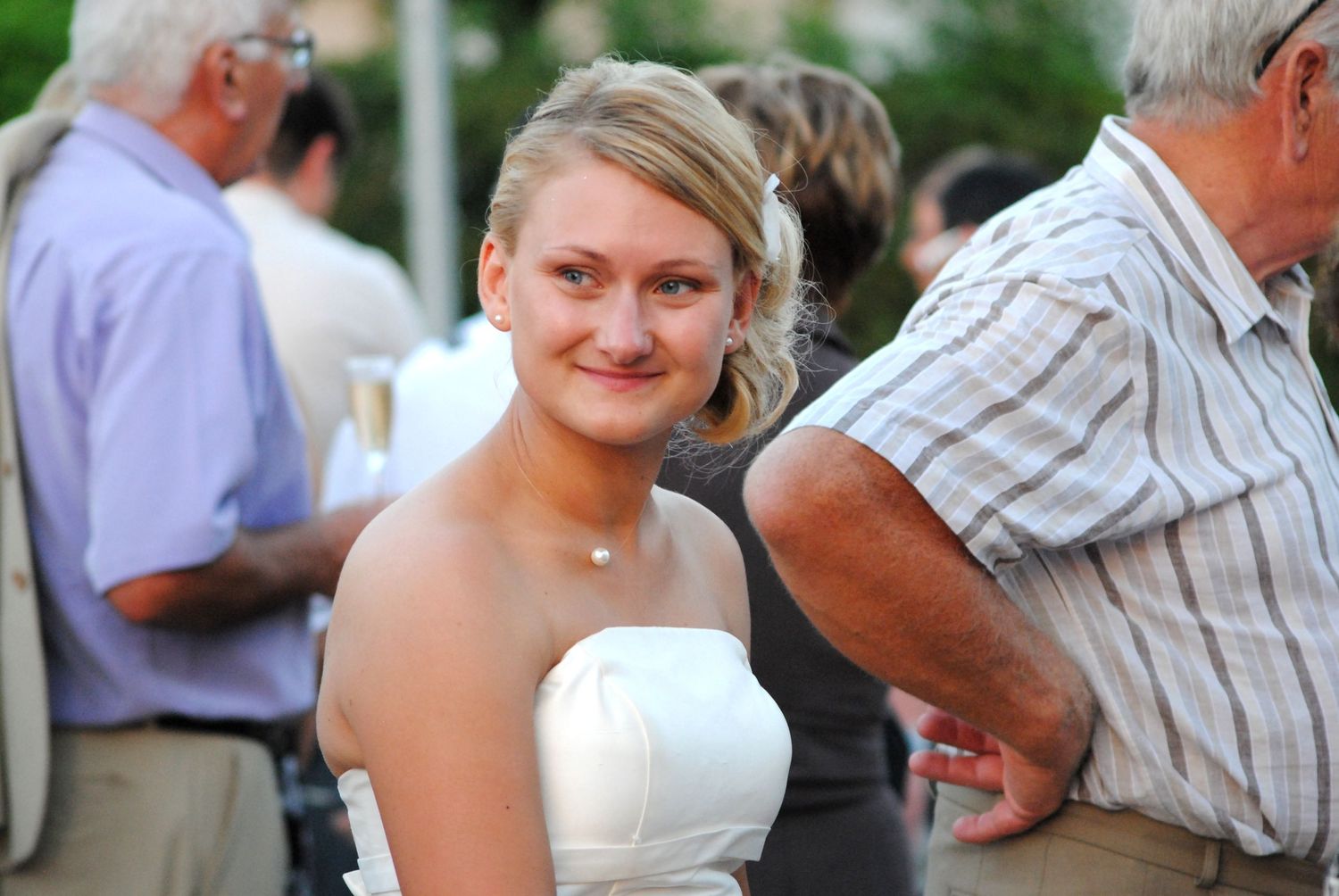 Young blonde woman in a white strapless dress smiling at an outdoor social gathering event.