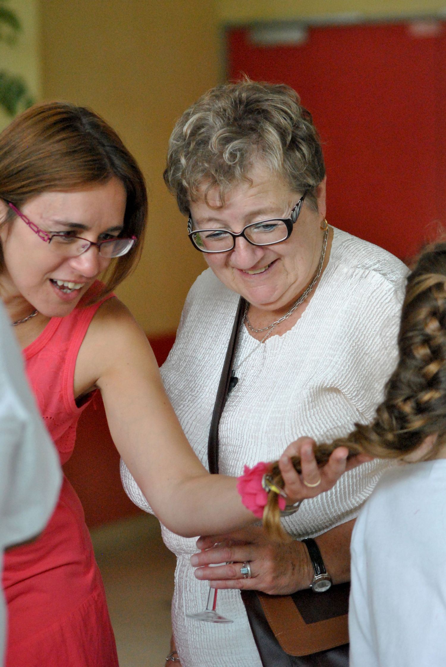Two women with glasses smile while examining a pink flower at a wedding reception.