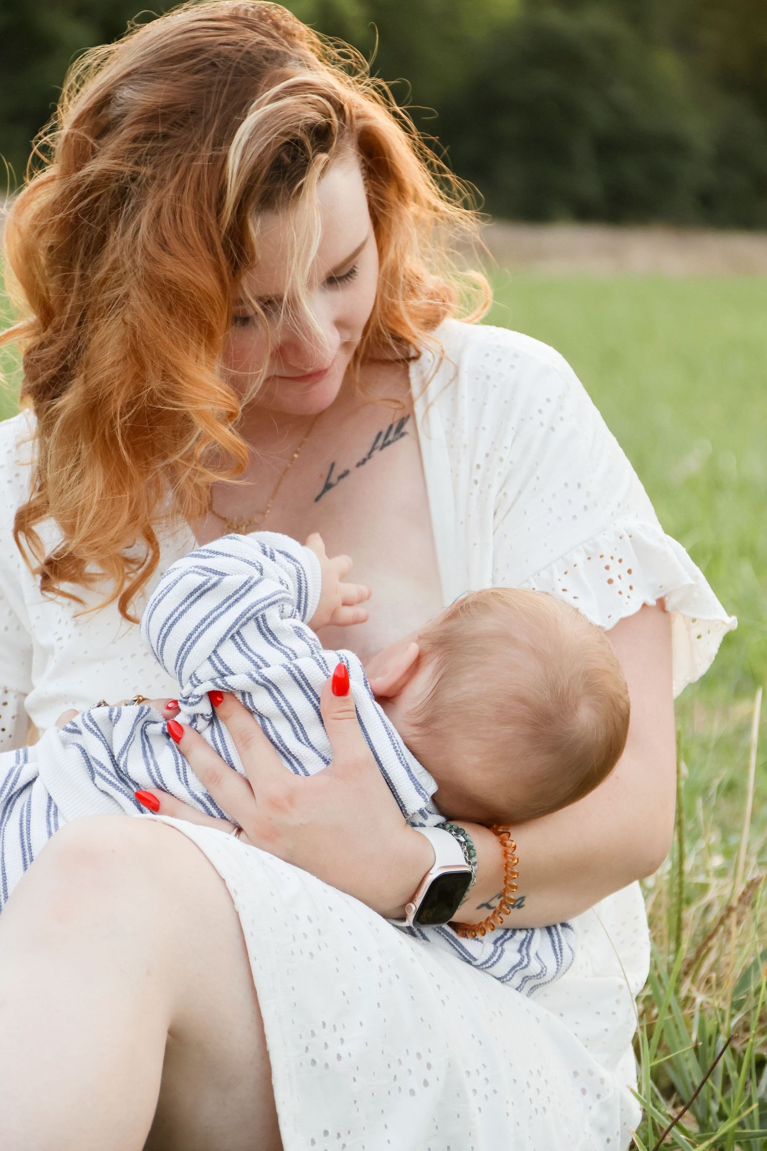 Redhead mother breastfeeding infant baby while sitting in green outdoor field.