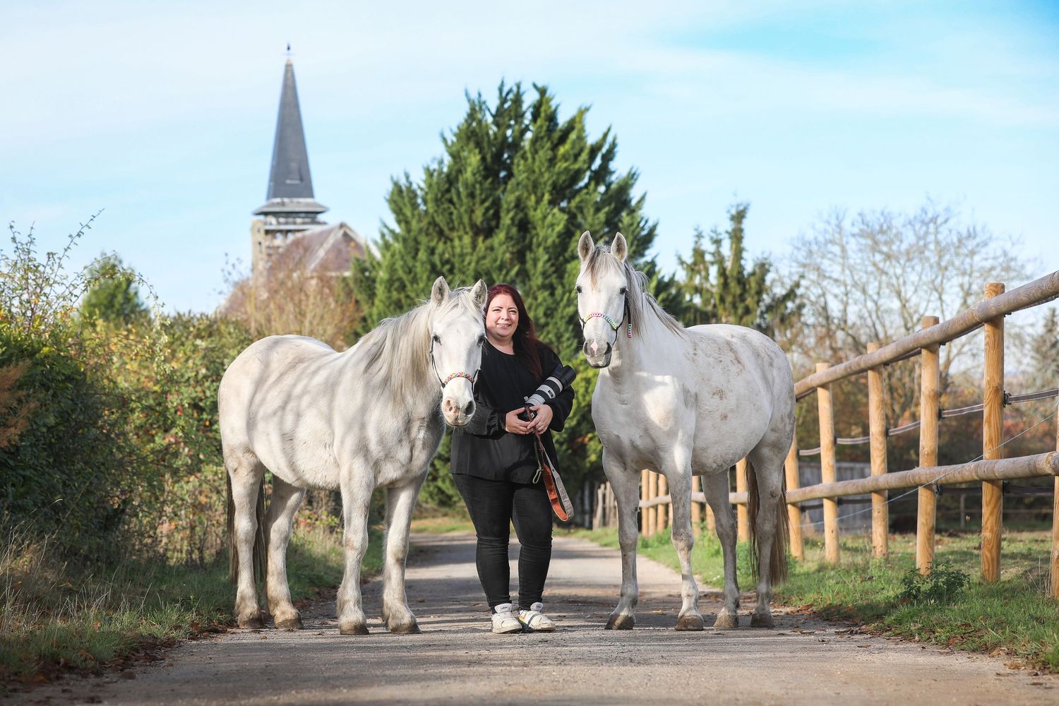 Woman walking between two white horses on rural path with church steeple visible in background.