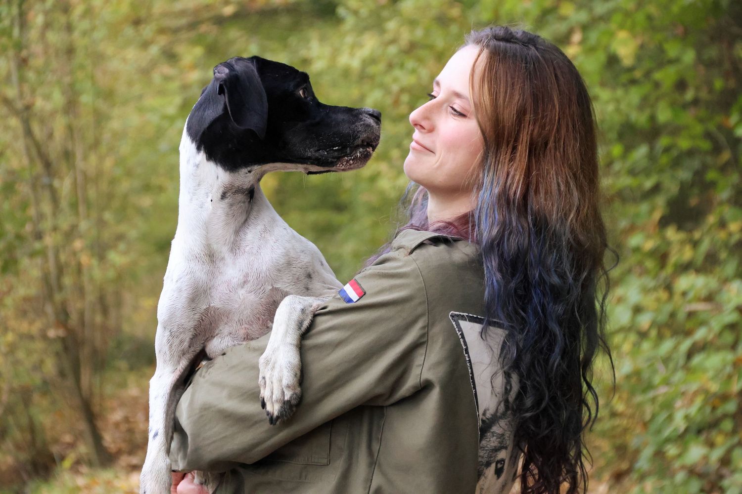 Woman holding black and white dog in autumn forest setting.