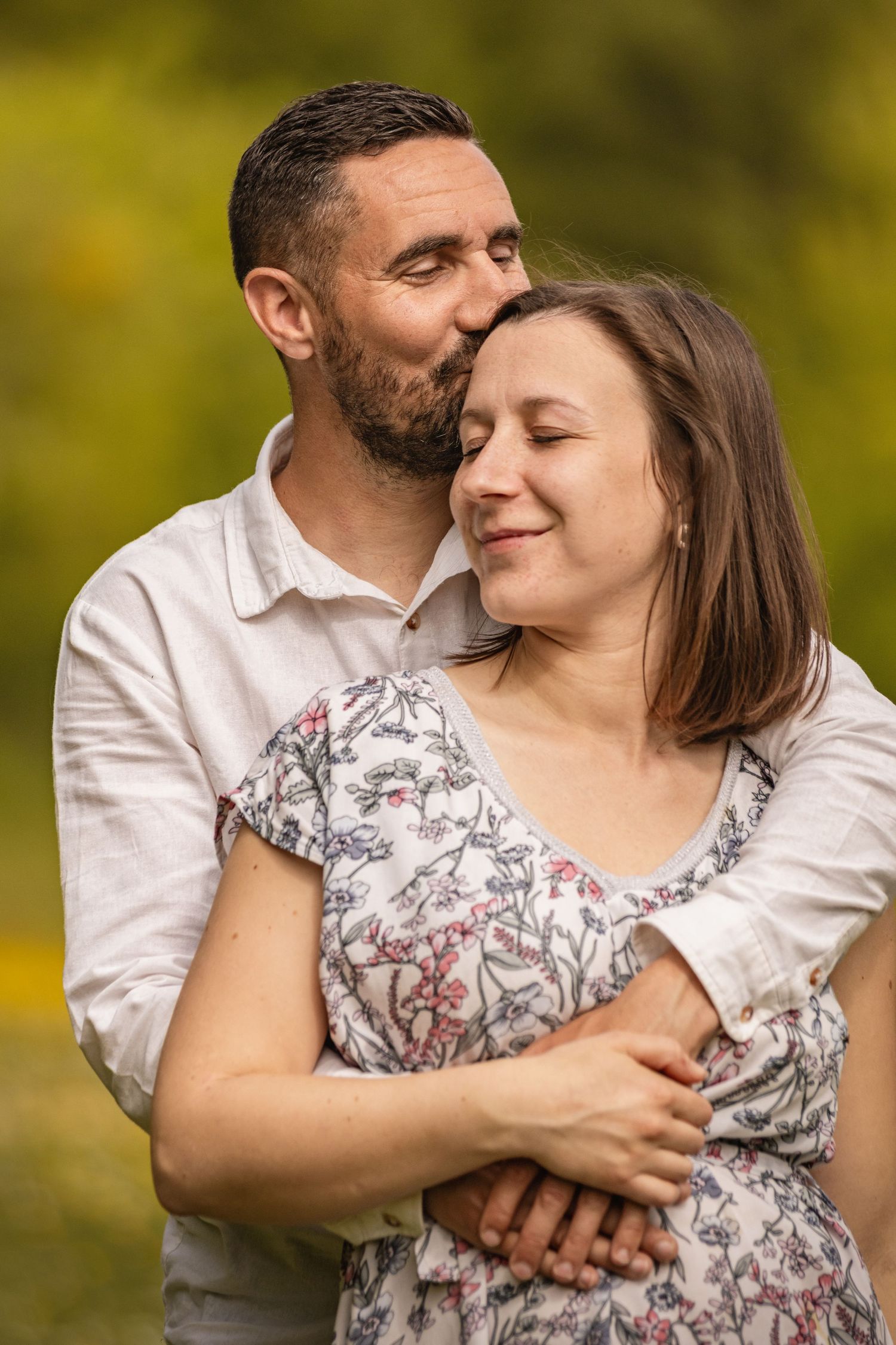 A bearded man tenderly kisses a smiling woman on the cheek while embracing her outdoors.