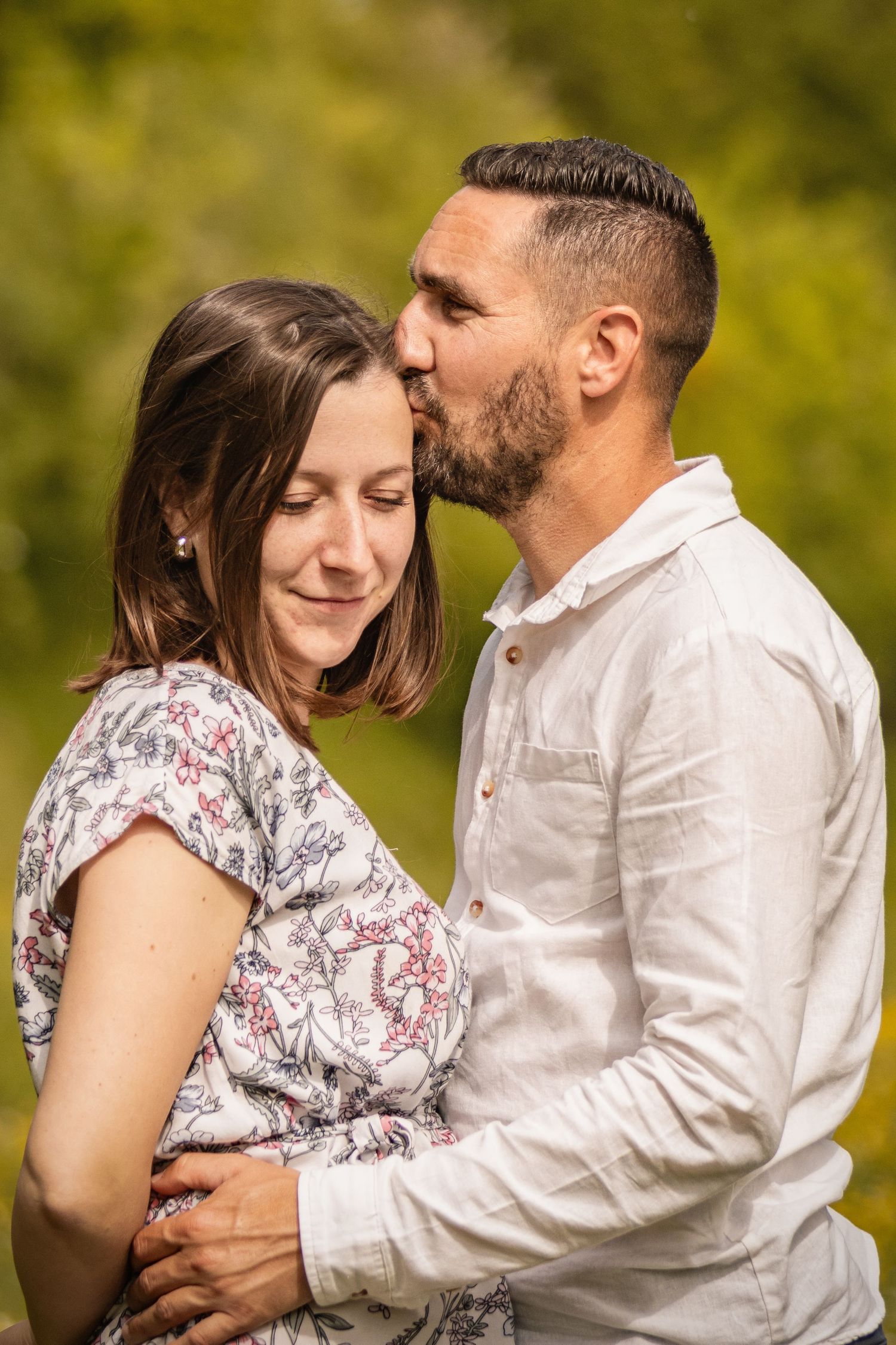 A bearded man in a white shirt tenderly kisses a smiling woman in a floral dress outdoors.